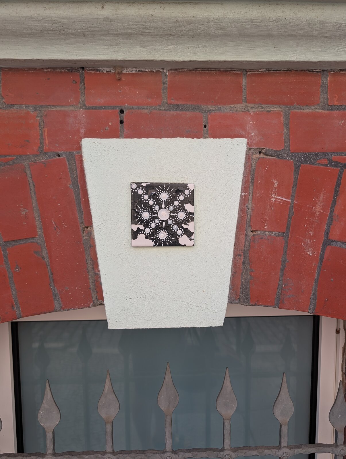 Eine kleine quadratische Keramikfliese ist an einer weiß gestrichenen Betonfläche zwischen roten Backsteinen angebracht. Die Fliese zeigt ein symmetrisches Mandala-Muster aus weißen Punkten unterschiedlicher Größe auf schwarzem und rosafarbenem Untergrund