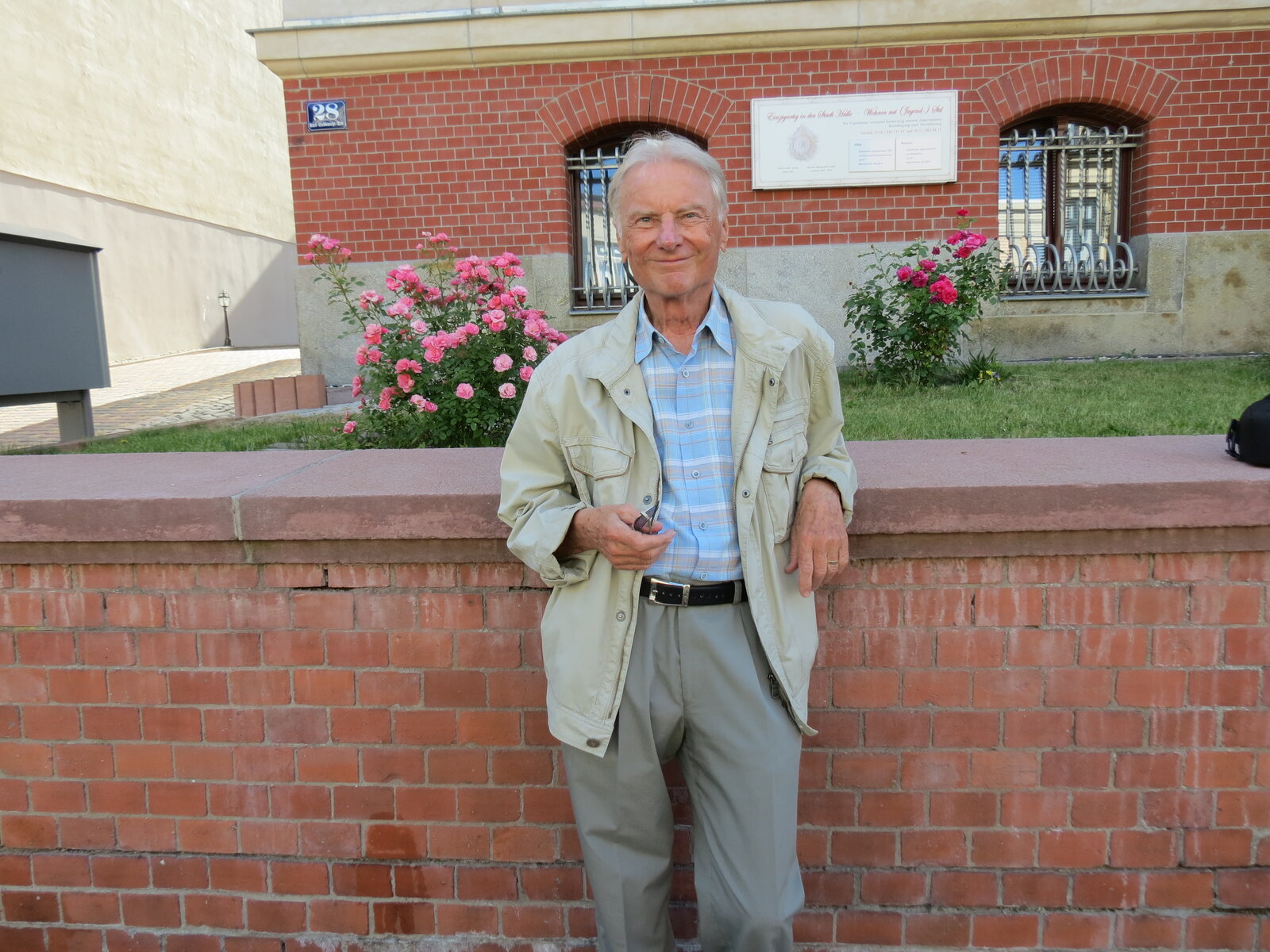 Ein älterer Mann in beigefarbener Jacke und kariertem Hemd steht vor einer roten Backsteinmauer mit rosa blühenden Pflanzen. Im Hintergrund ist ein Wohngebäude mit Fenstern und weißem Dach zu sehen.