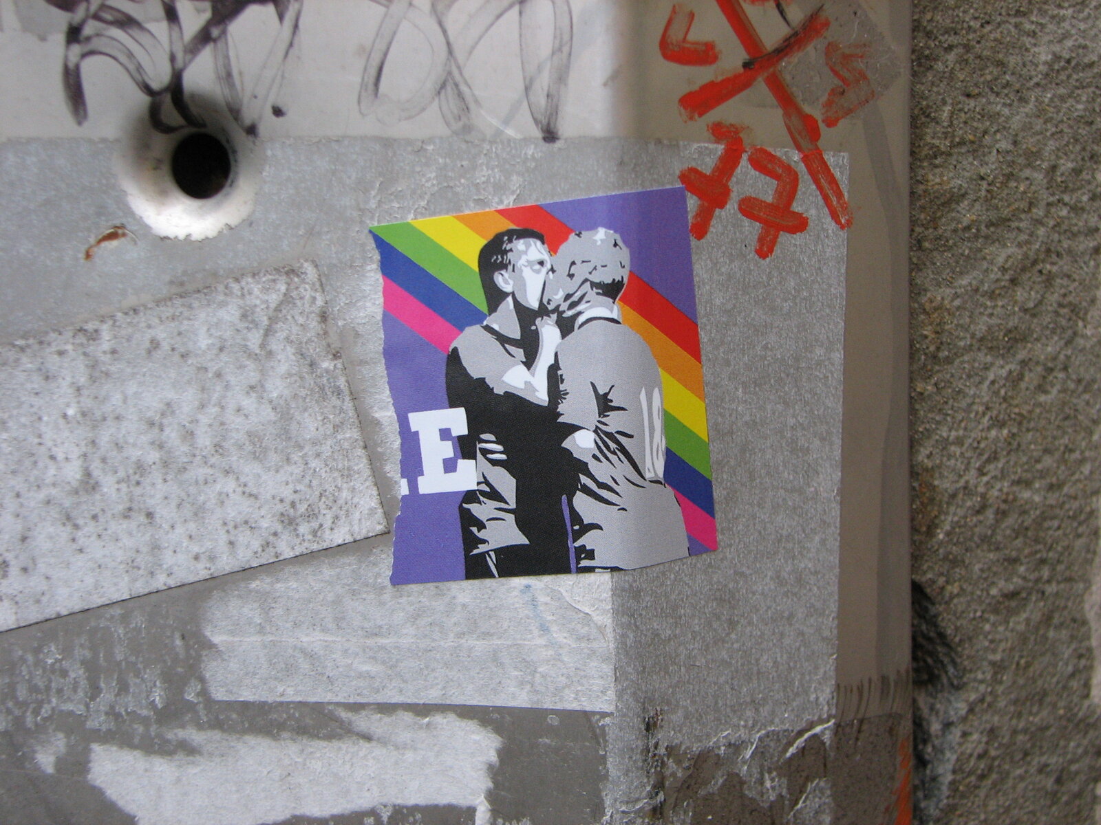 Ein Sticker auf grauer Wand zeigt zwei Personen in Schwarz-Weiß vor einem bunten Regenbogen-Hintergrund mit Blau-, Rot- und Gelbtönen.