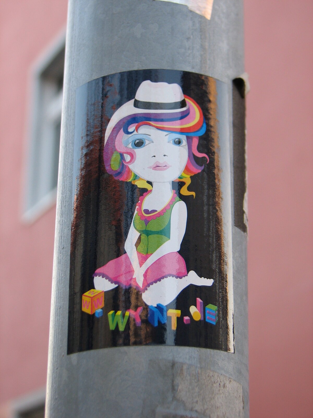 Aufgeklebter farbiger Sticker auf grauer Metallröhre vor rosafarbenem Hintergrund. Das Motiv zeigt eine stilisierte Figur mit mehrfarbig gelockertem Haar, Hut, großem Auge und bunter Kleidung. Unten im Bild ist ein farbiger Schriftzug zu sehen.