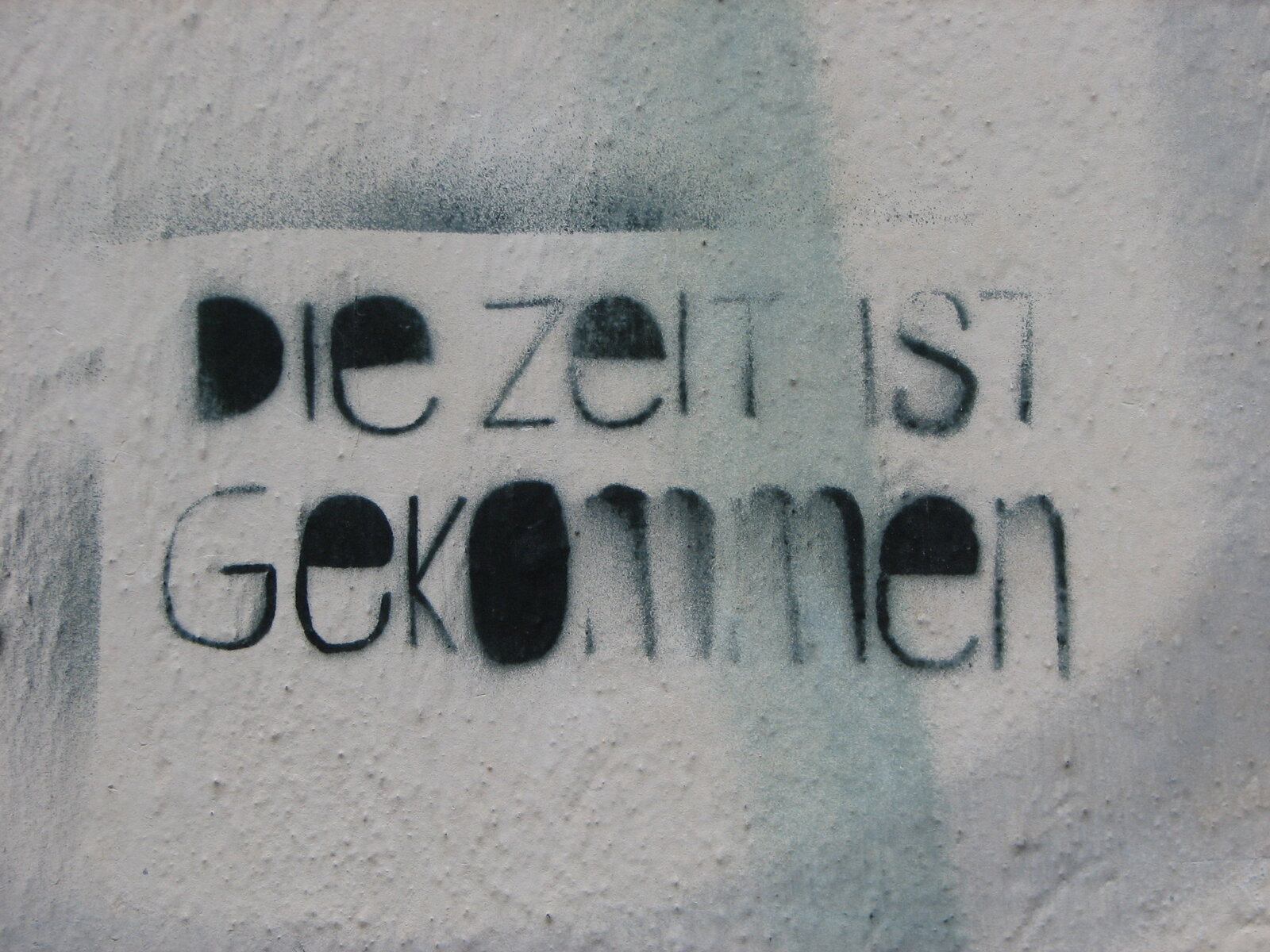 Schwarze Schriftzug-Graffiti auf hellgrauer Wand mit teilweise verblasstem oder übermaltem Hintergrund. Der Text "die zeit ist gekommen" ist in einfachen, geometrischen Buchstaben geschrieben.