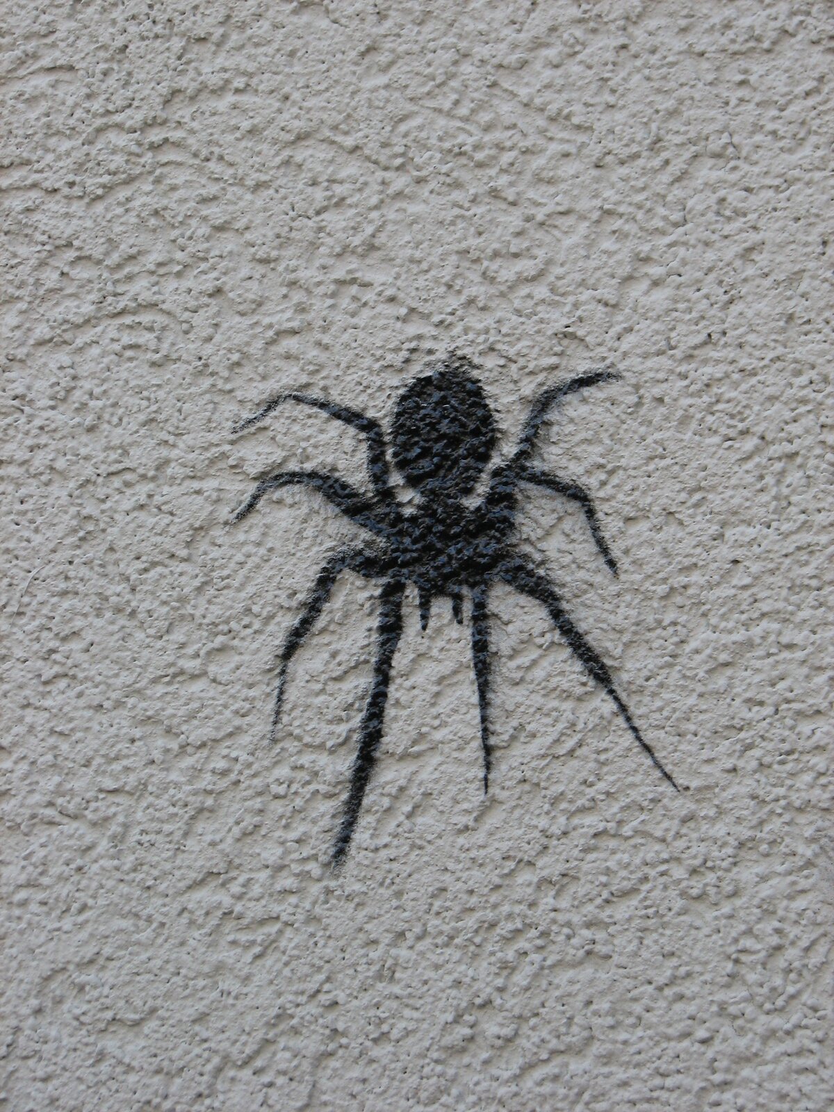 Stencil-Schablonendruck einer schwarzen Spinne auf grauem Wandgrund. Die Figur zeigt ein symmetrisches Spinnenmotiv mit ausgebreiteten Beinen und zentralem Körper.
