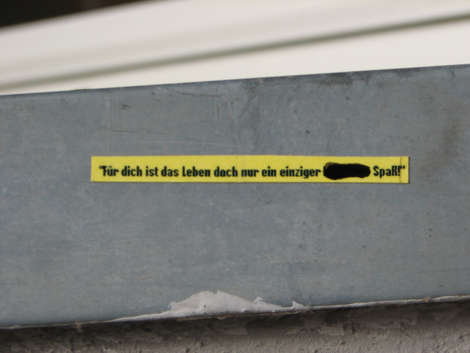 Ein gelber Streifen mit schwarzer Schrift auf grauer Betonwand. Der Text "Für dich ist das leben doch nur ein einziger ... Spaß" ist teilweise durch eine schwarze Markierung unlesbar gemacht.