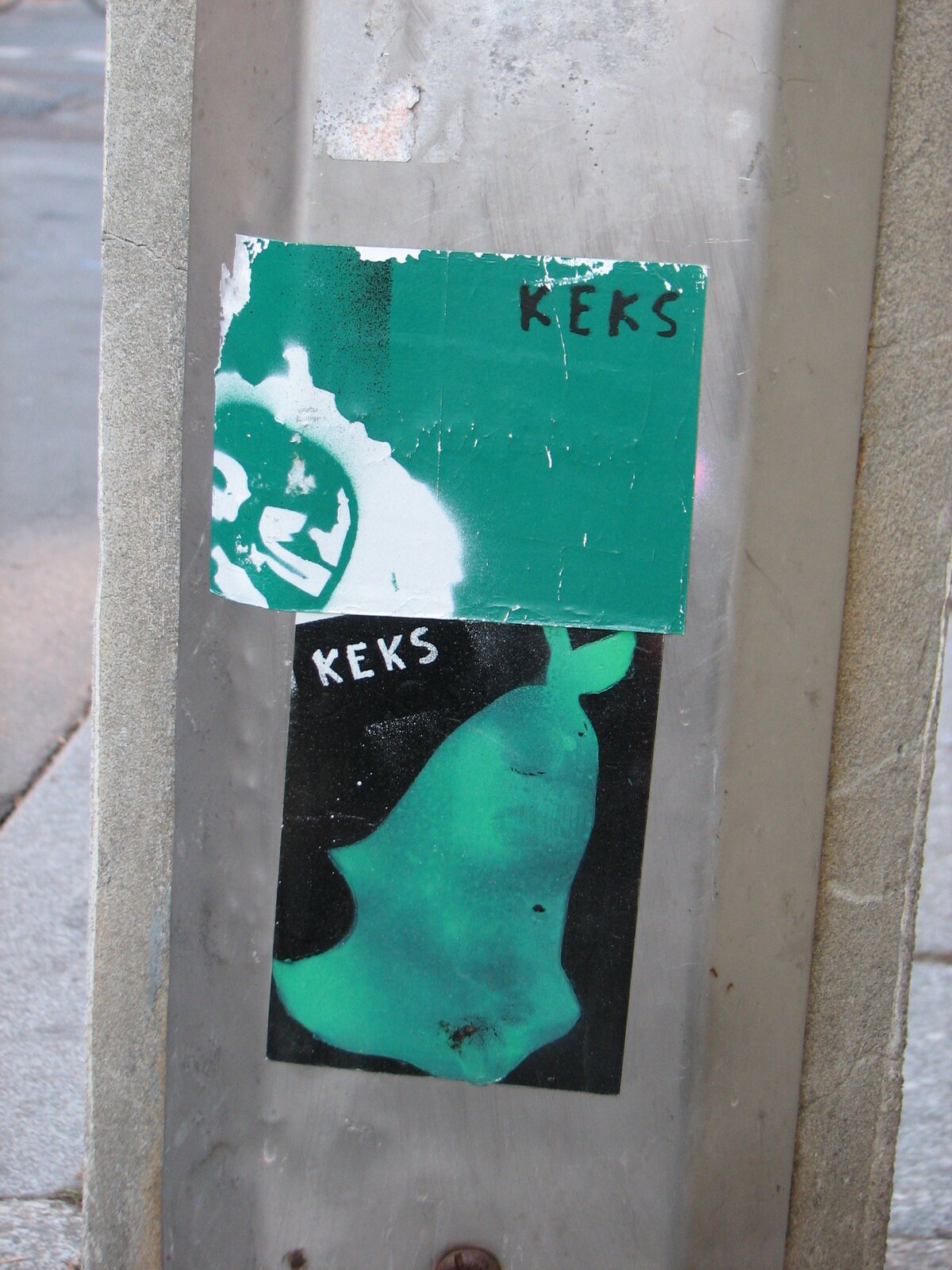 Zwei übereinander angebrachte Sticker auf grauem Untergrund: oben ein grünes Sticker-Fragment mit weißem Motiv und Schriftzug, unten ein schwarzes Sticker-Rechteck mit grünem Farbverlauf und Text.