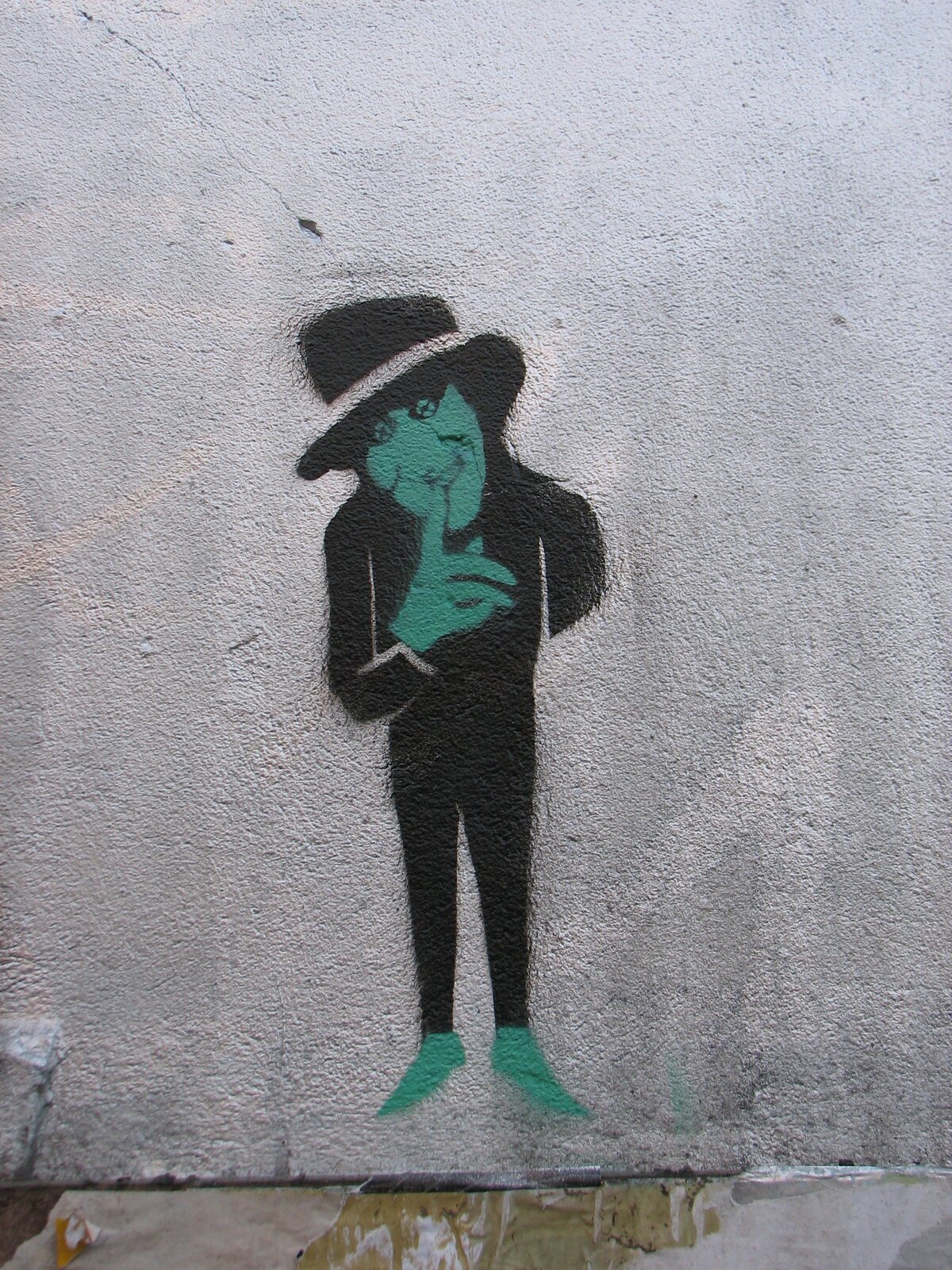Sprühmalerei auf grauer Wand: Eine schlanke, schwarz umrissene Figur in Anzug und Hut mit grünem Gesicht und grünen Beinen, Hand nachdenklich ans Kinn gelegt.