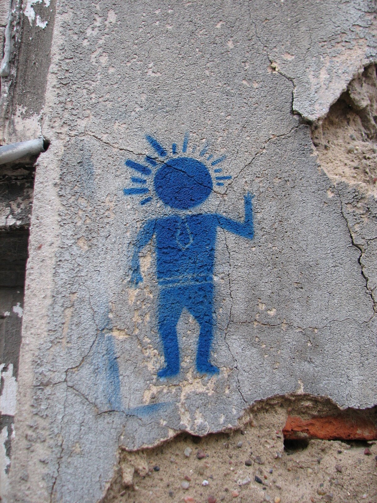 Blaues Stencil-Graffito auf grauer Betonwand: eine stilisierte menschliche Figur mit strahlenförmigem Kopf, die eine Hand hebt. Die Wand ist verwittert und weist Risse sowie Feuchtigkeitsspuren auf.