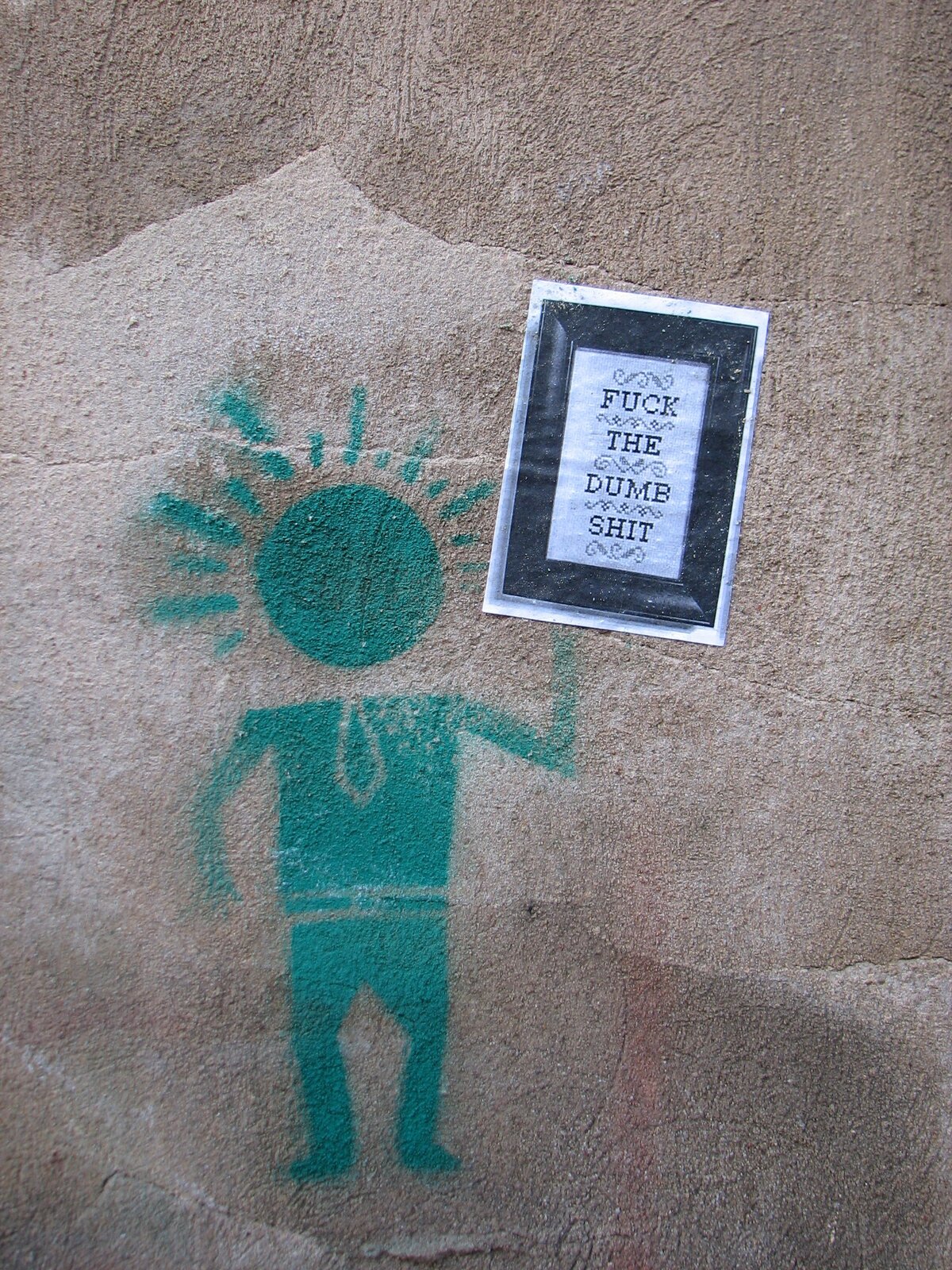 Türkis-grüne Stencil-Figur mit ausgebreiteten Armen und strahlenförmigem Kopf auf grauer Betonwand. Daneben ein rechteckiger gerahmter Sticker mit Text: Fuck the dumb shit.