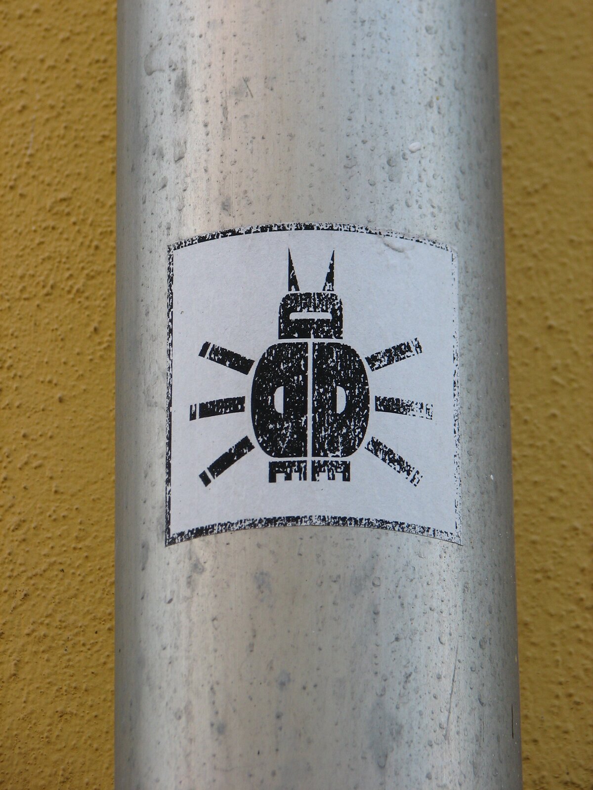 Auf einem verwitterten metallischen Rohr ist ein schwarz-weißer Sticker mit einem Motiv einer Marienkäfer-artigen Figur mit zwei aufragenden Spitzen angebracht. Das Motiv ist von strahlenförmigen Linien umgeben.