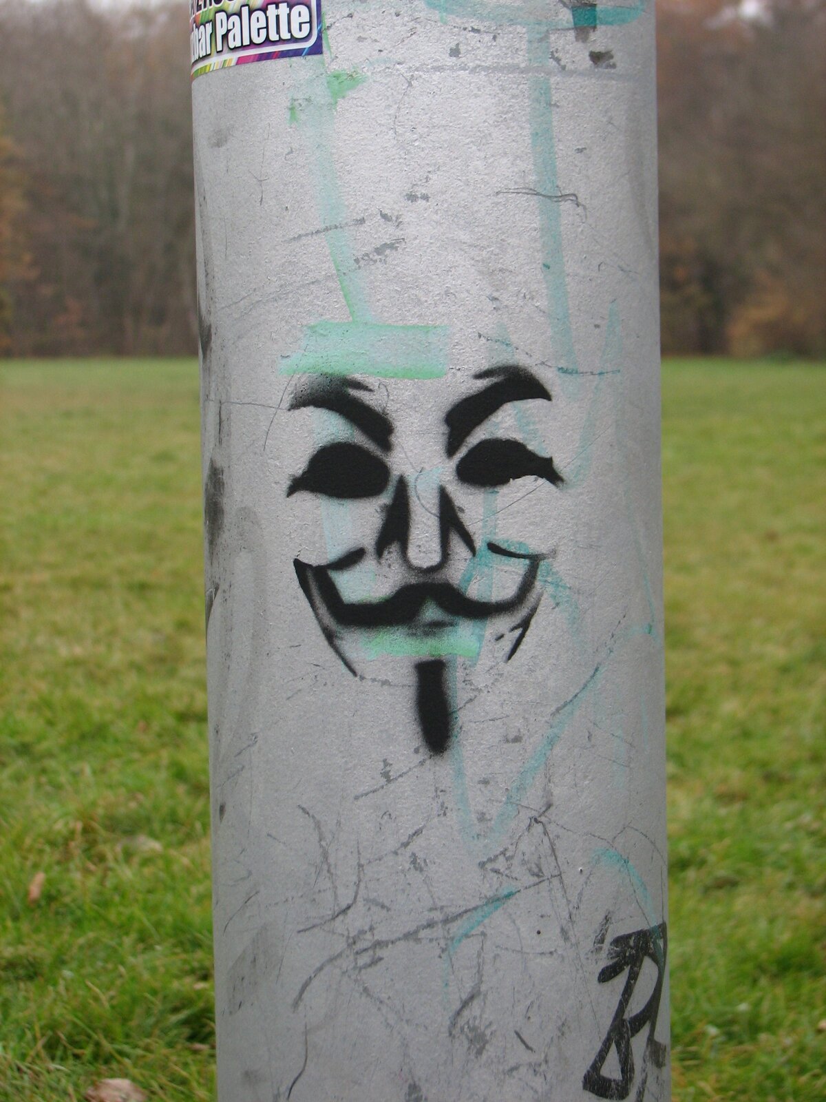 Stencil-Porträt eines Gesichts mit mandelförmigen Augen, breiter Nase und Schnurrbart auf einer grauen Metallröhre. Schwarze Schablonenschrift, grüne Markierungen und Kratzer im Untergrund sichtbar.