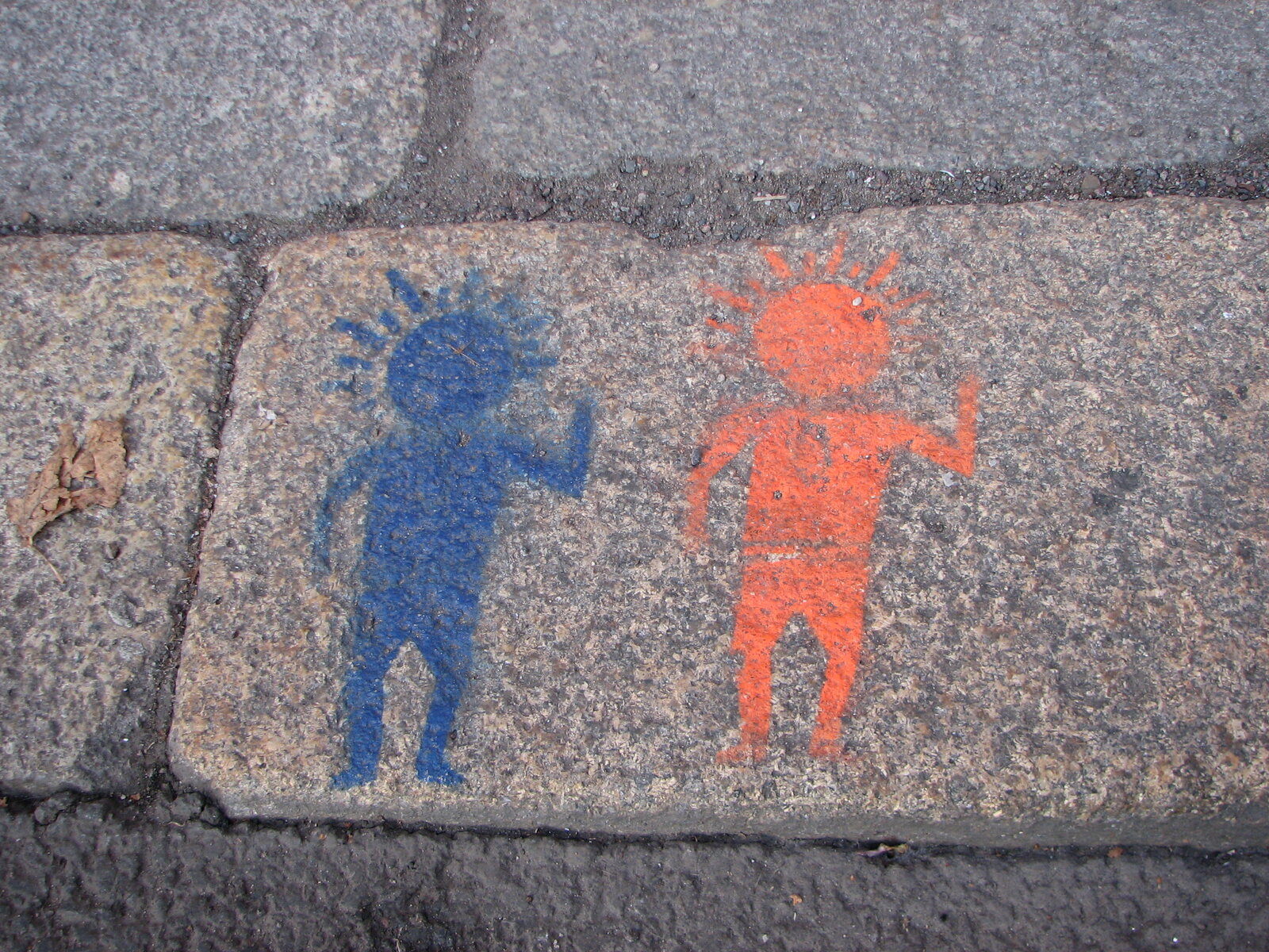 Zwei einfache, stilisierte menschliche Figuren in Stencil-Technik auf grauem Pflasterstein: eine blaue Figur mit erhobenem Arm links, eine rote Figur mit erhobenem Arm rechts.
