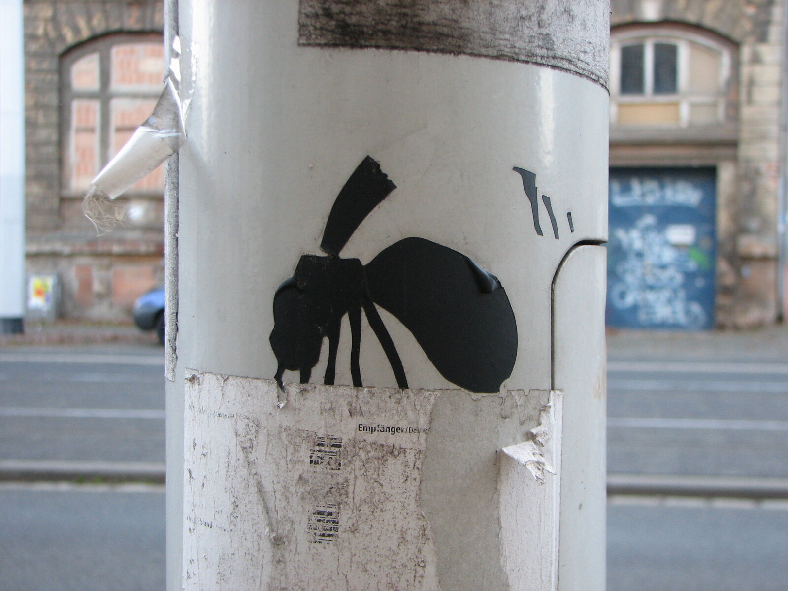 Schwarze Scherenschnitt-Silhouette einer Hornisse auf weißem Untergrund an einem Stromkasten oder Pfeiler im urbanen Raum. Im Hintergrund Backsteingebäude und Straße.