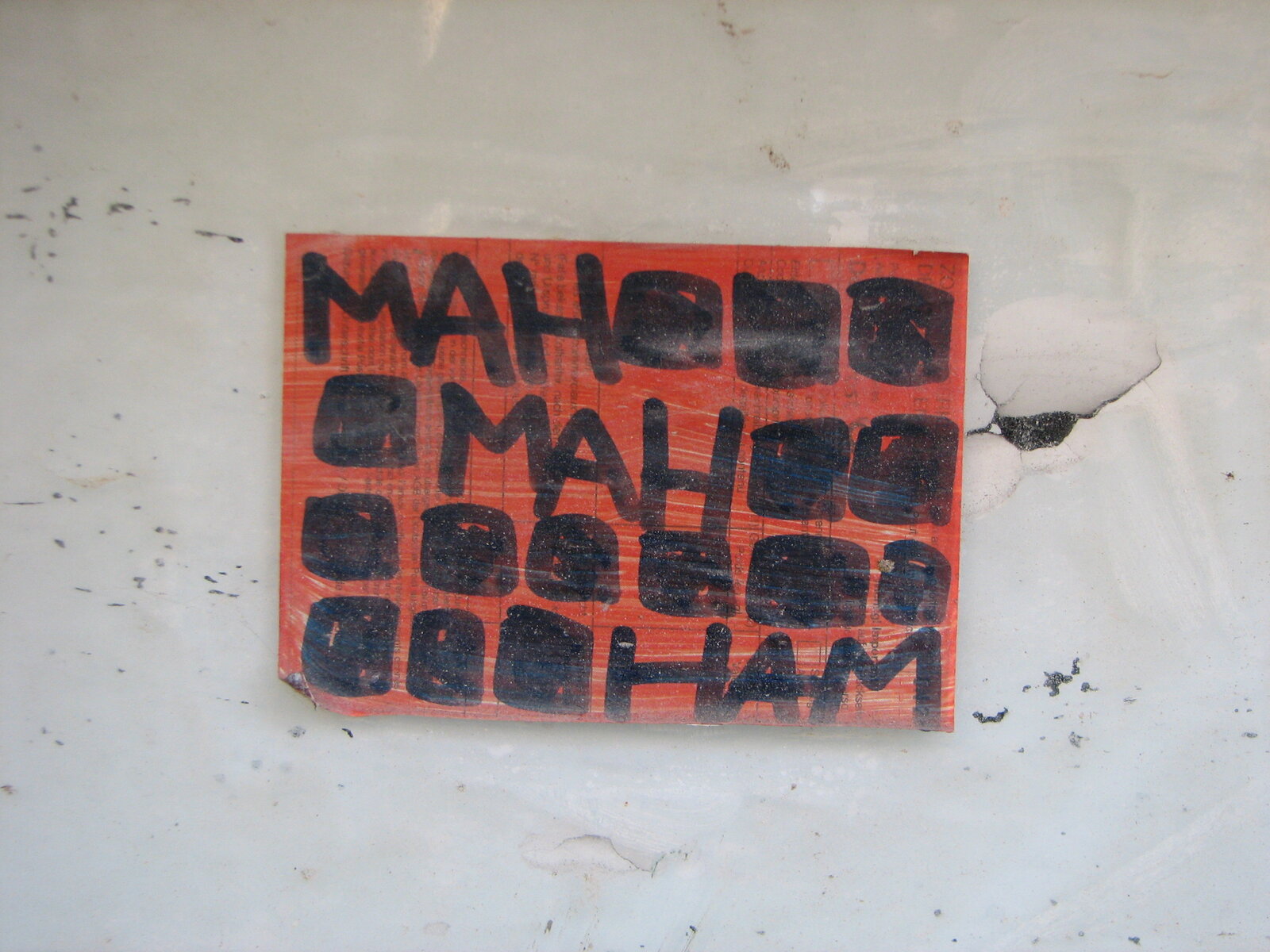 Ein rotes Papier-Sticker auf weißer Wand mit schwarzem Text in mehreren Reihen, teilweise überlagert oder unlesbar gemacht durch schwarze Rechtecke.