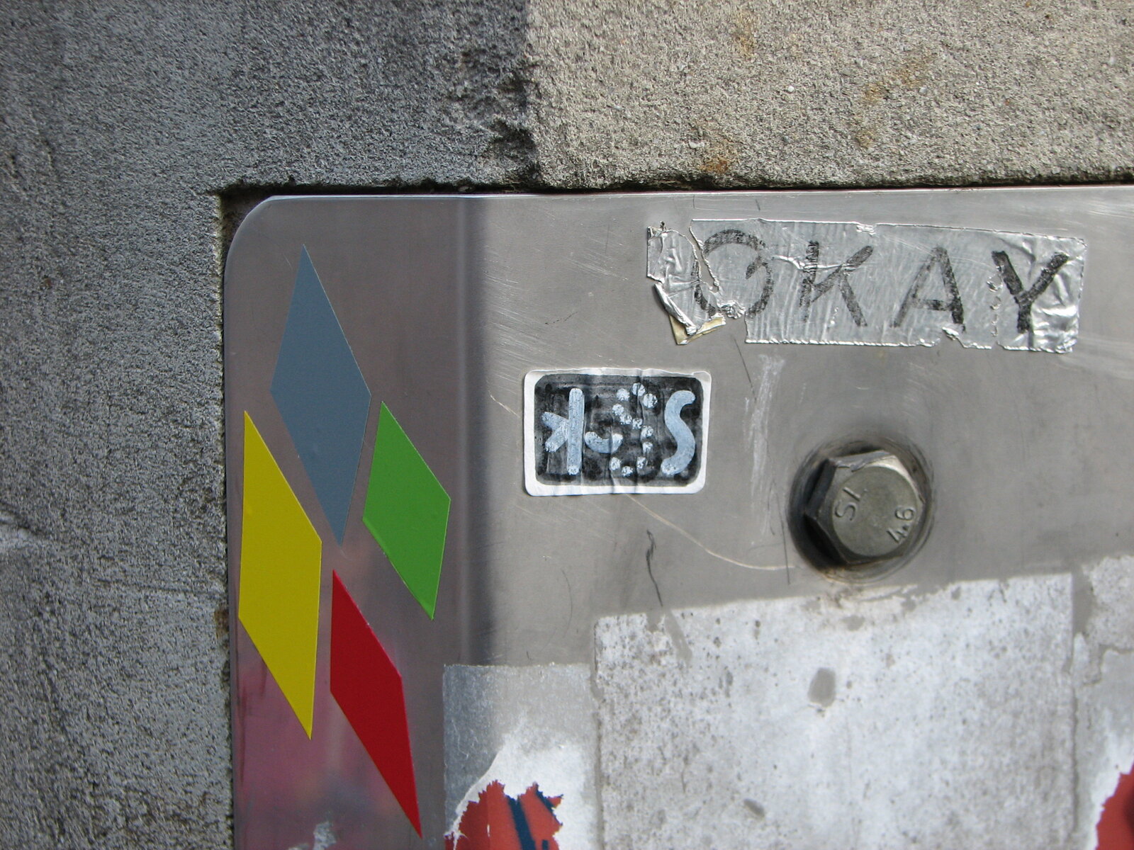 Aufgeklebter Sticker auf grauer Metallfläche mit geometrischen Formen in Blau, Grün, Gelb und Rot, daneben eine weiße Sticker-Box mit schwarzem Muster und ein rundes Metallnietnagel.