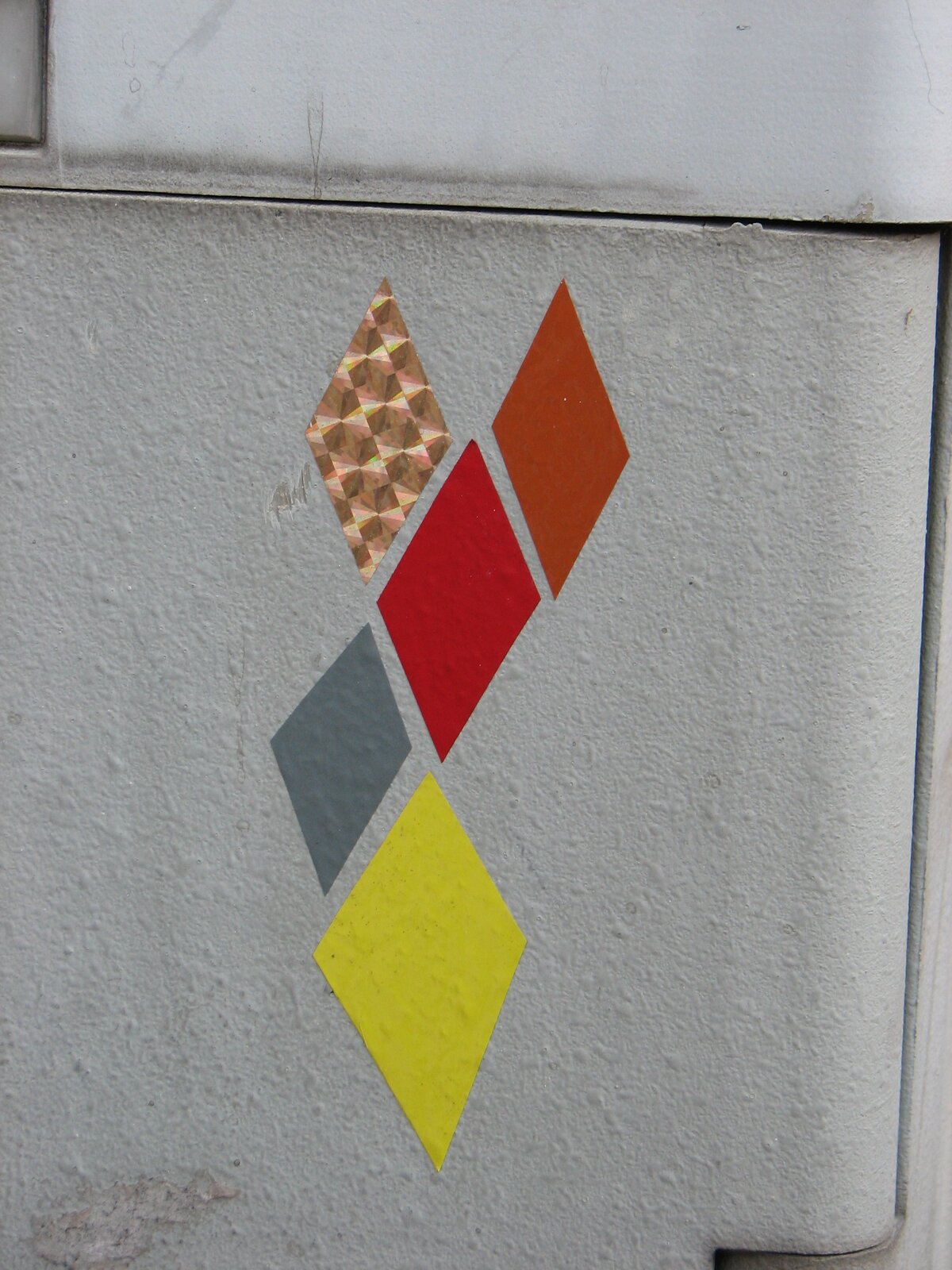 Vier diamantförmige Papierstücke in Creme-Weiß mit Punktmuster, Rot, Grau und Gelb sind diagonal übereinander auf einer weißen Fliesenwand geklebt.