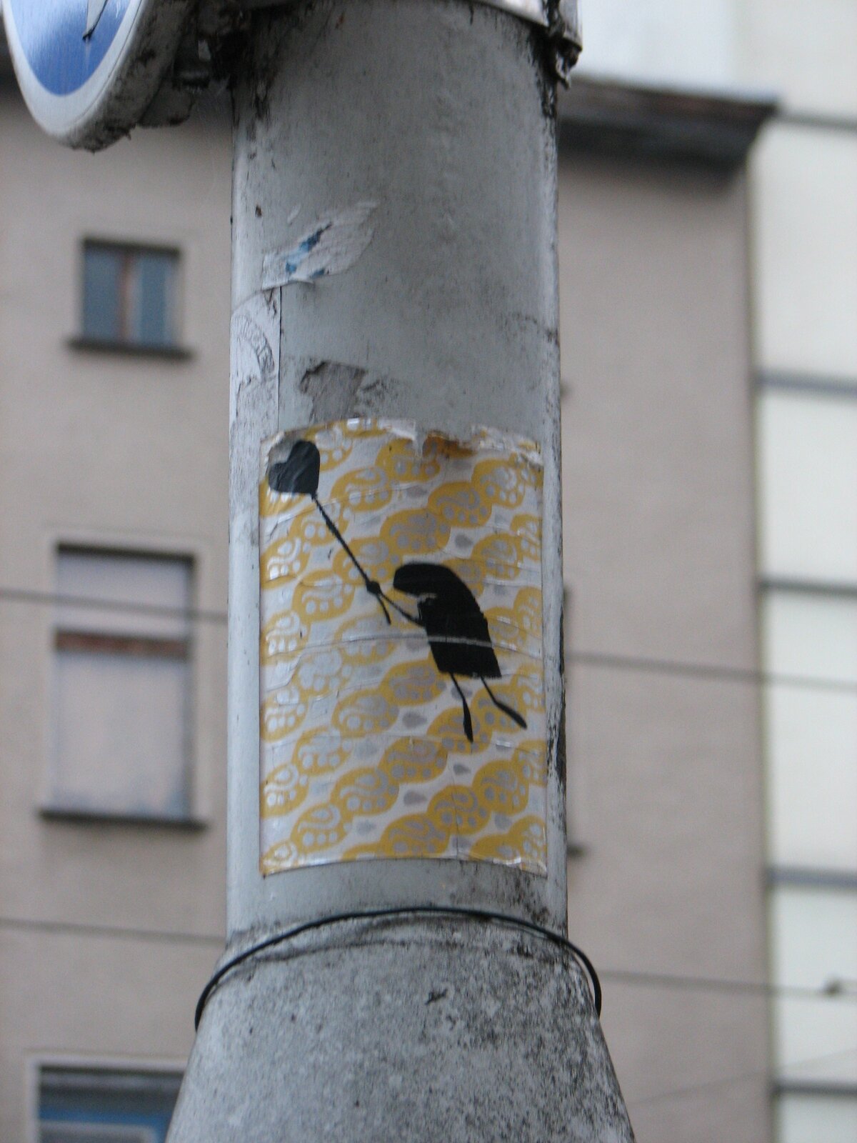 Ein Sticker auf einem grauen Metallpfosten zeigt eine stilisierte Figur, die mit einem Luftballon abhebt. Der Hintergrund ist gelb und hat ein geometrisches Muster. Der Pfosten steht vor einem mehrstöckigen Wohngebäude.