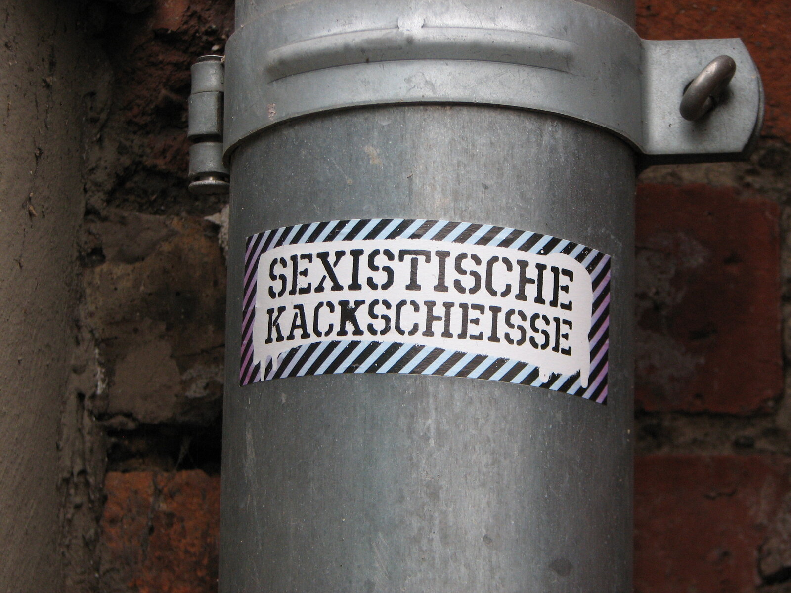 Ein grauer Metallzylinder oder Rohrabschnitt mit aufgeklebtem rechteckigem Sticker. Der schwarzweiß gestreifte Sticker trägt in großen Majuskeln die Aufschrift "SEXISTISCHE KACKSSCHEISSE".