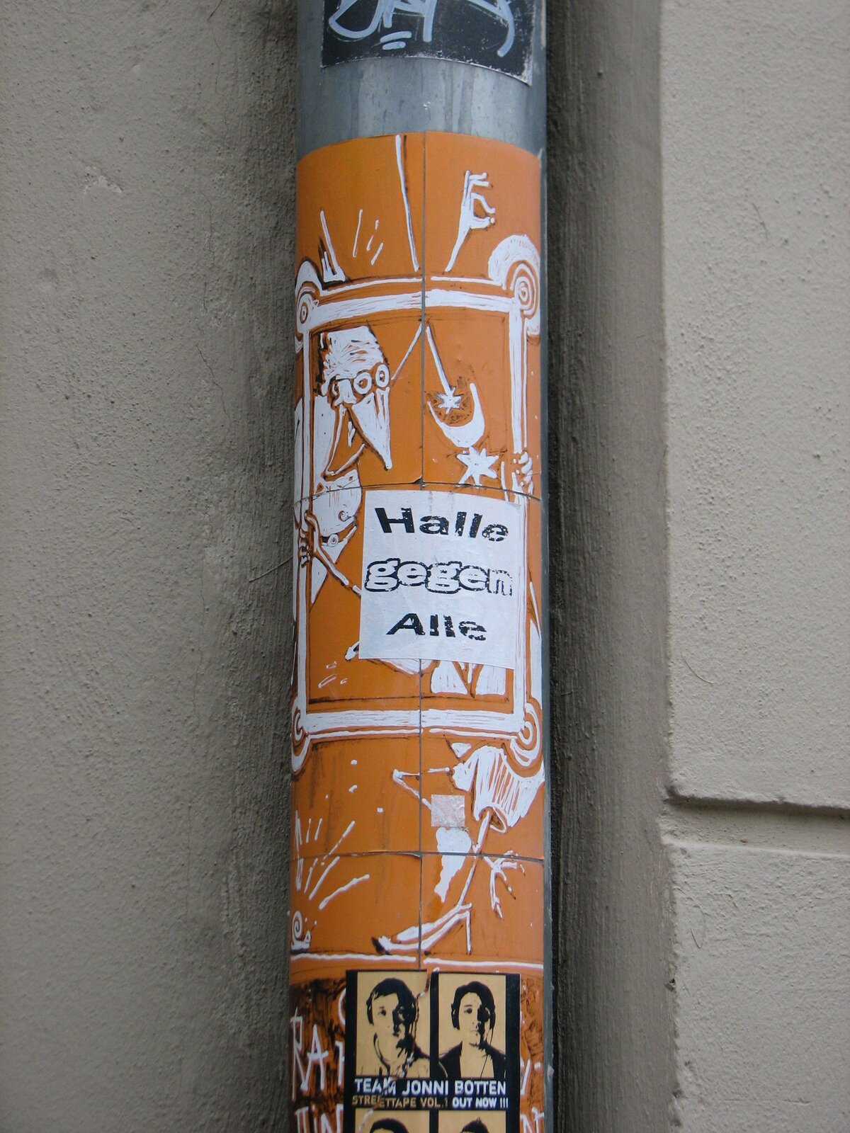 Ein oranges Sticker-Motiv auf einem runden Metallrohr zeigt schematische Figuren und Szenen. Darauf ein Papiersticker mit Schriftzug "HALLE GEGEN ALLE" und weiteren Abbildungen.
