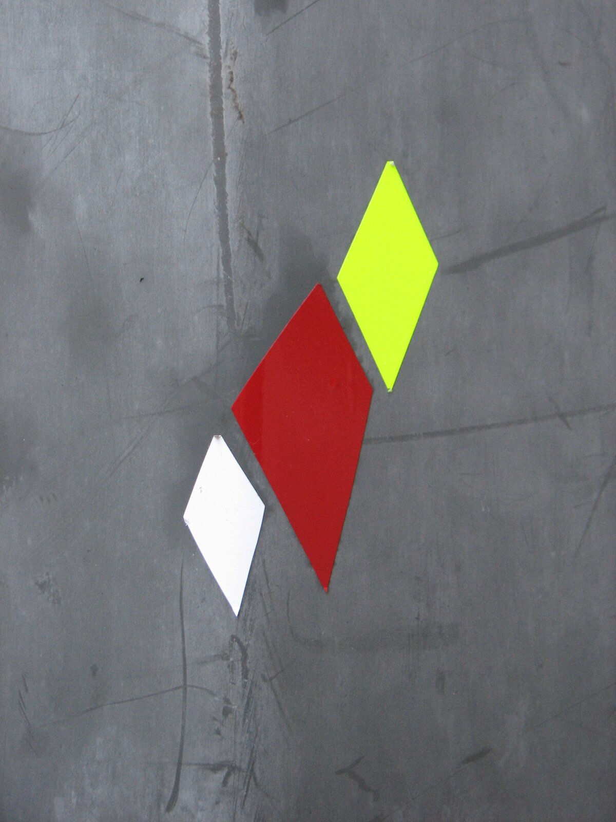 Drei geometrische Diamantformen in Weiß, Rot und Neongelb kleben vertikal versetzt auf einer grauen Betonwand.