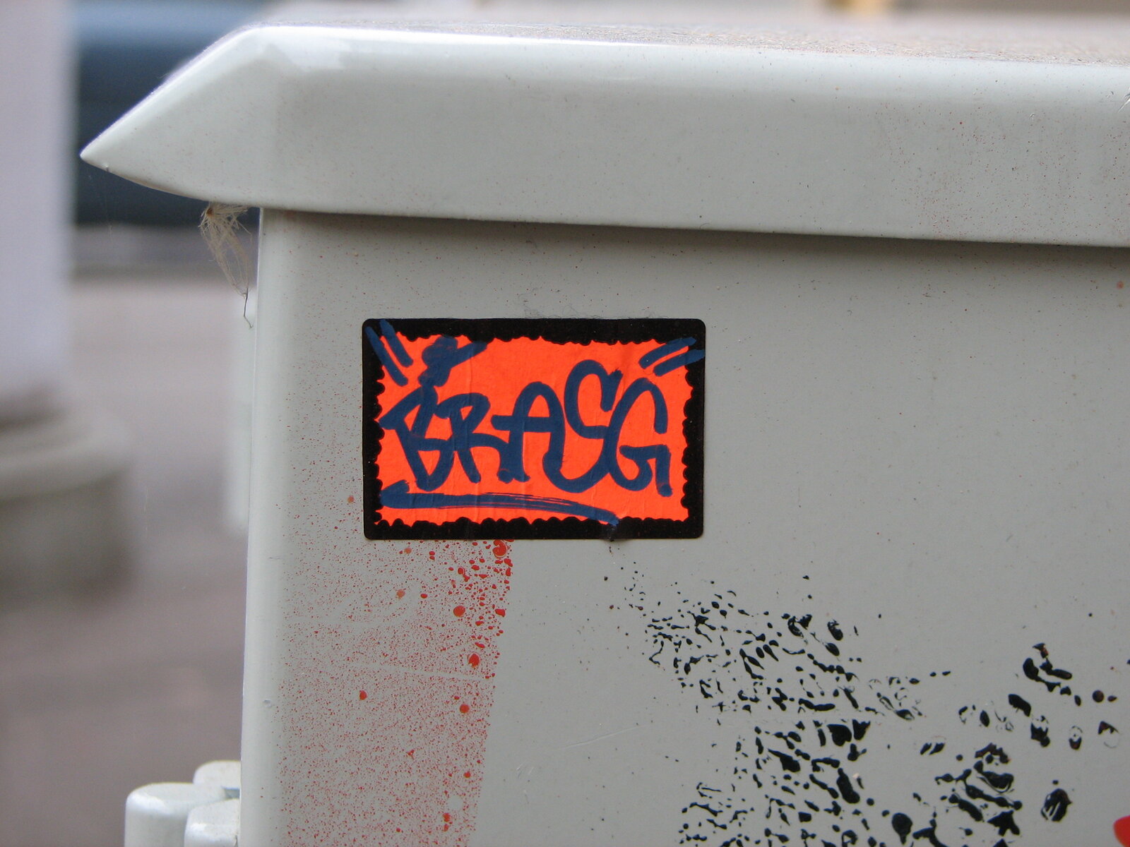 Ein leuchtend orange-rotes Sticker-Motiv mit schwarzem Rand auf weißer Oberfläche. Das Tag zeigt stilisierte, verschlungene Lettern in klassischem Graffiti-Stil. Darunter sind rote und schwarze Sprühspuren und Farbabdrücke sichtbar.