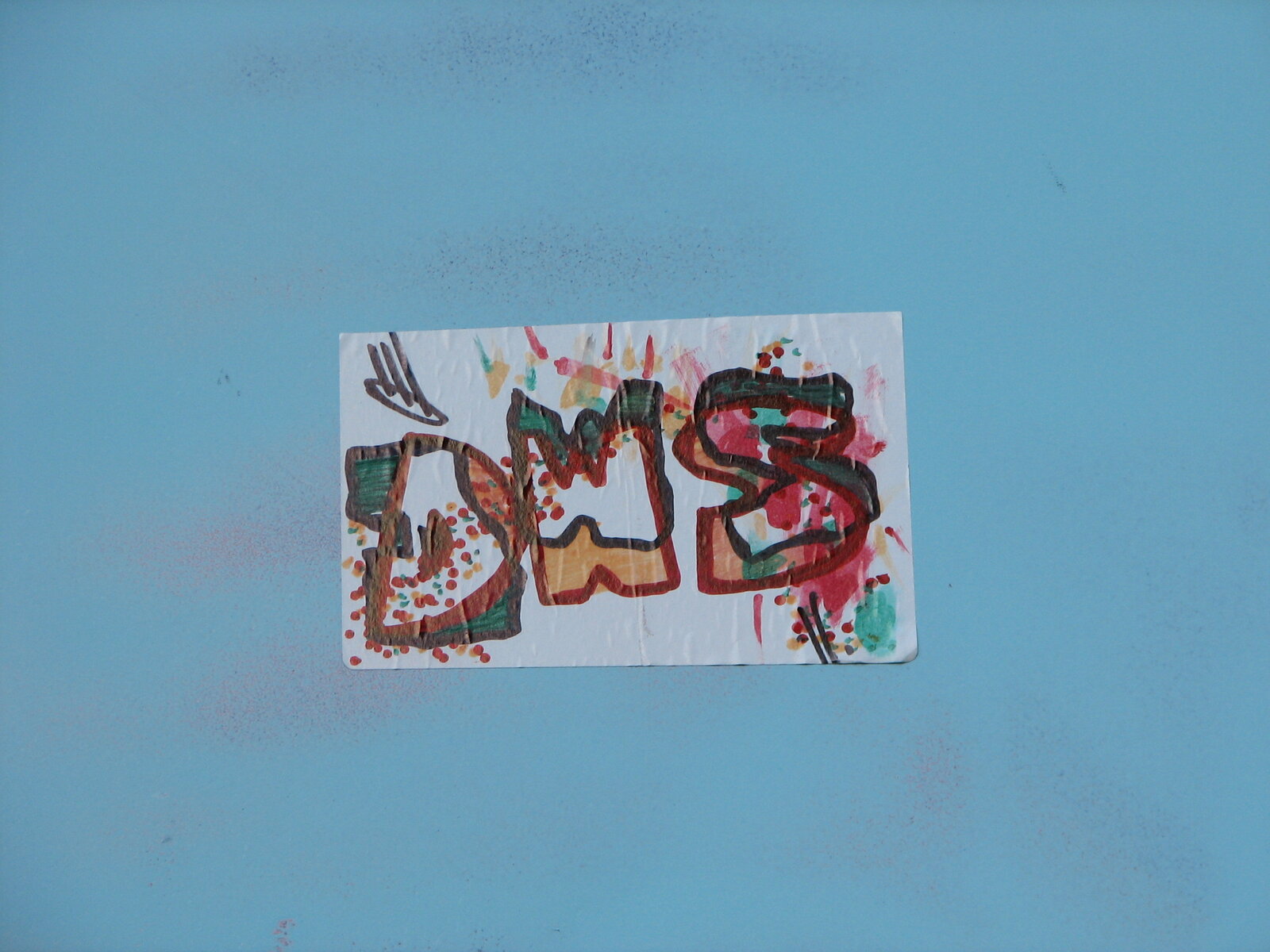 Ein rechteckiger Sticker auf hellblauem Untergrund zeigt abstrakte Buchstaben in Rot, Grün, Gelb und Schwarz mit dynamischen Formen und Overlays; kleine dekorative Elemente umgeben die Komposition.