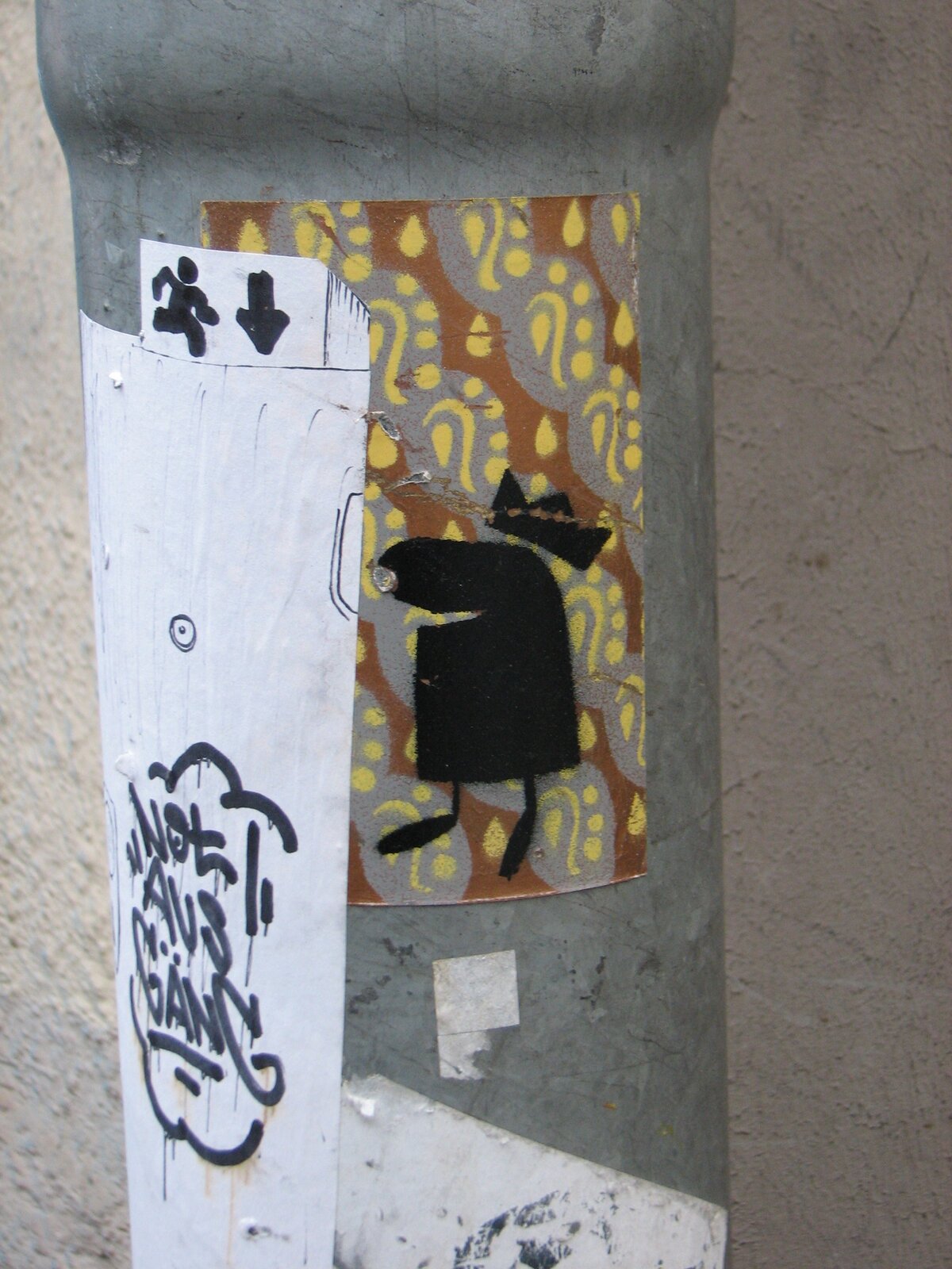 An einem grauen Betonrohr sind zwei Street-Art-Werke angebracht: links ein weißer Sticker mit schwarzen Pfeilen und einem Tag, rechts ein farbiges Papier-Cut-out oder Sticker mit gelben und rötlichen Mustern und einer schwarzen Figurendarstellung.