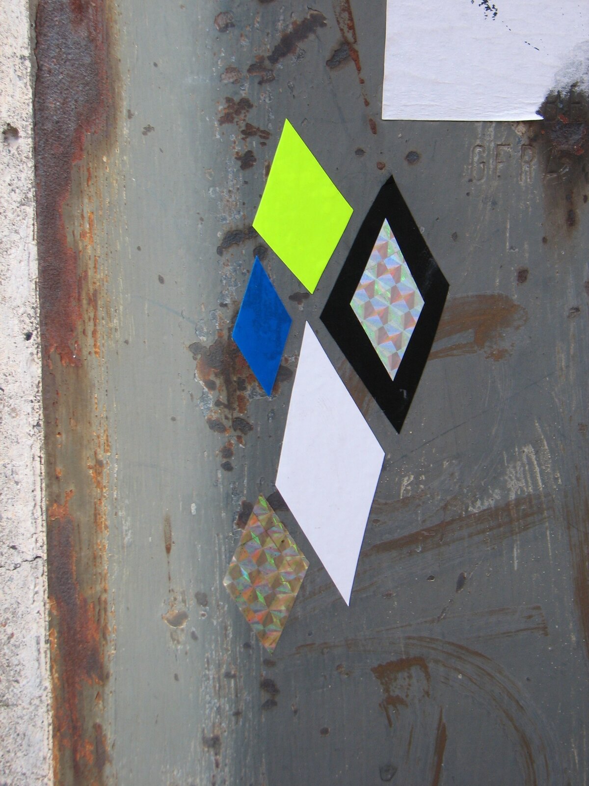 Mehrfarbige geometrische Diamanten-Cut-outs auf verwitterter Metallfläche: Ein fluoreszierendes gelb-grünes Viereck, ein blaues Viereck, ein schwarzes Viereck mit Gitternetz-Muster, ein weißes Viereck und ein weiteres geometrisches Element mit Pastell-Ras