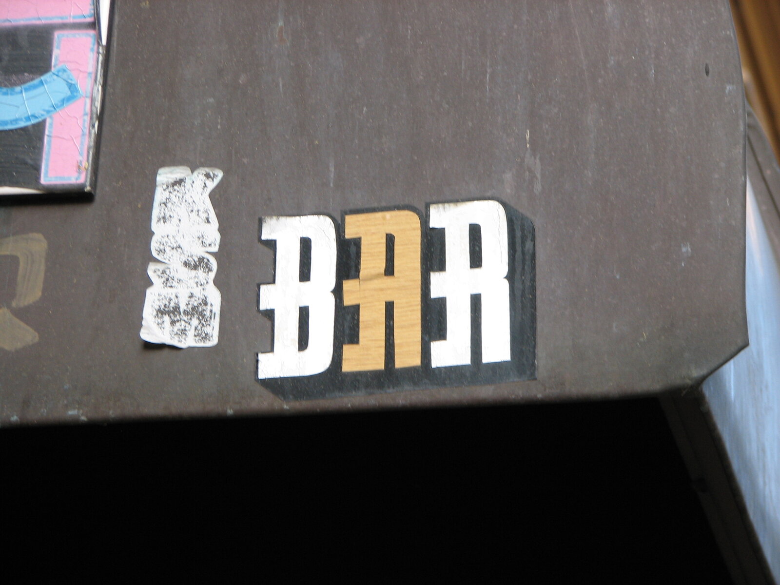 Aufgeklebter weißer und goldener Schriftzug "BAR" auf dunkelgrauer Oberfläche, links daneben ein teilweise abgelöster weißer Sticker mit Textur.