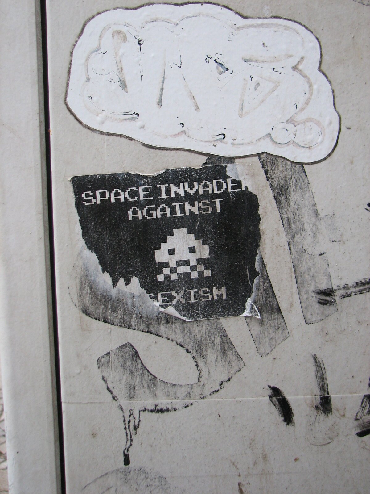 Sticker-Kunstwerk mit weißem, pixeligen Raumschiff-Motiv oben und darunter eine schwarze Fläche mit weißem Pixel-Art-Alien und Text in Großbuchstaben. Die Arbeit ist auf grauem Untergrund angebracht.