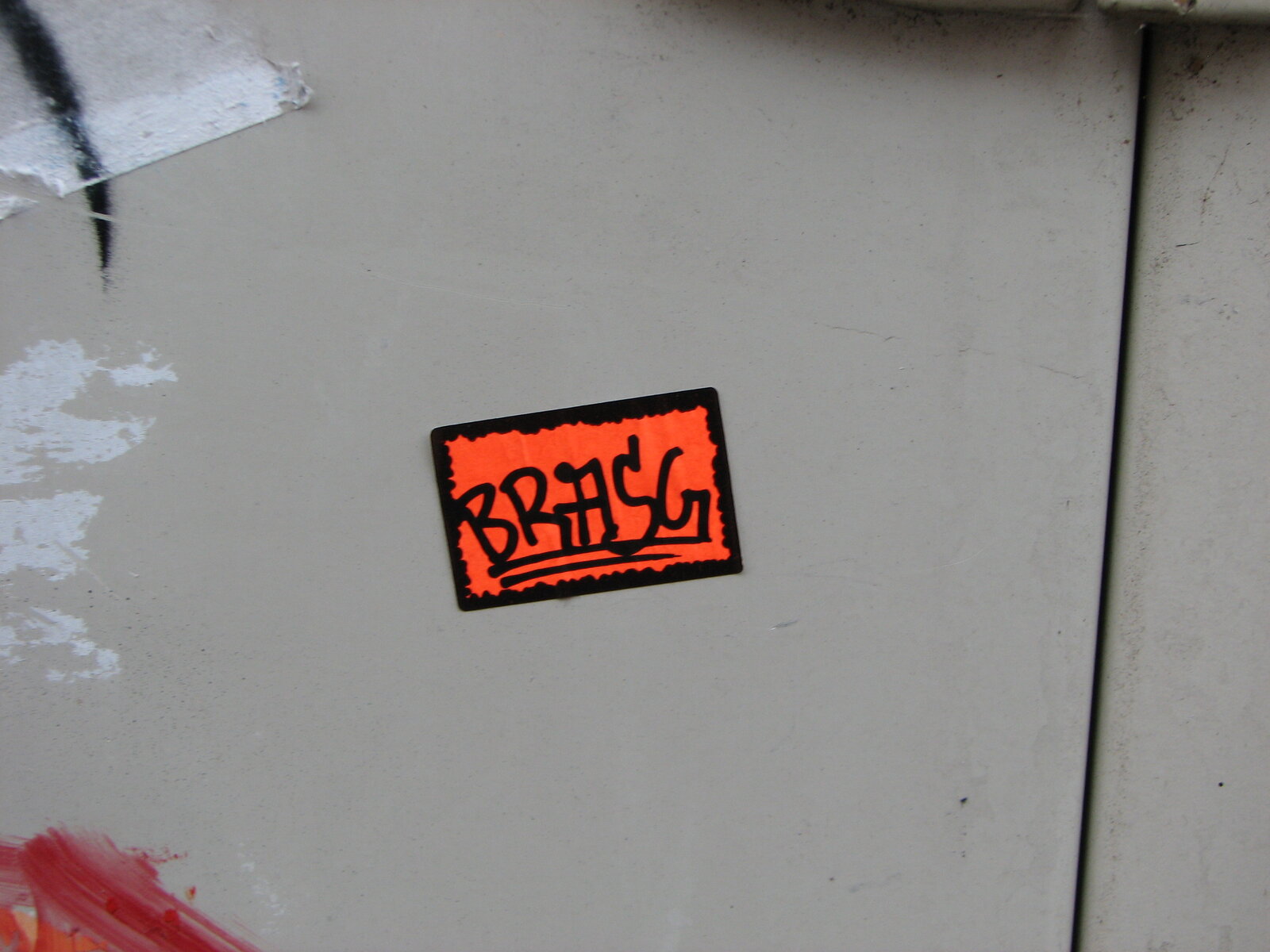 Ein rechteckiger orange-roter Sticker mit schwarzem Rand auf hellgrauer Wand. Der Sticker zeigt eine stilisierte Schrift in Schwarz mit zwei horizontalen Linien darunter. Zu lesen ist "BRASG".