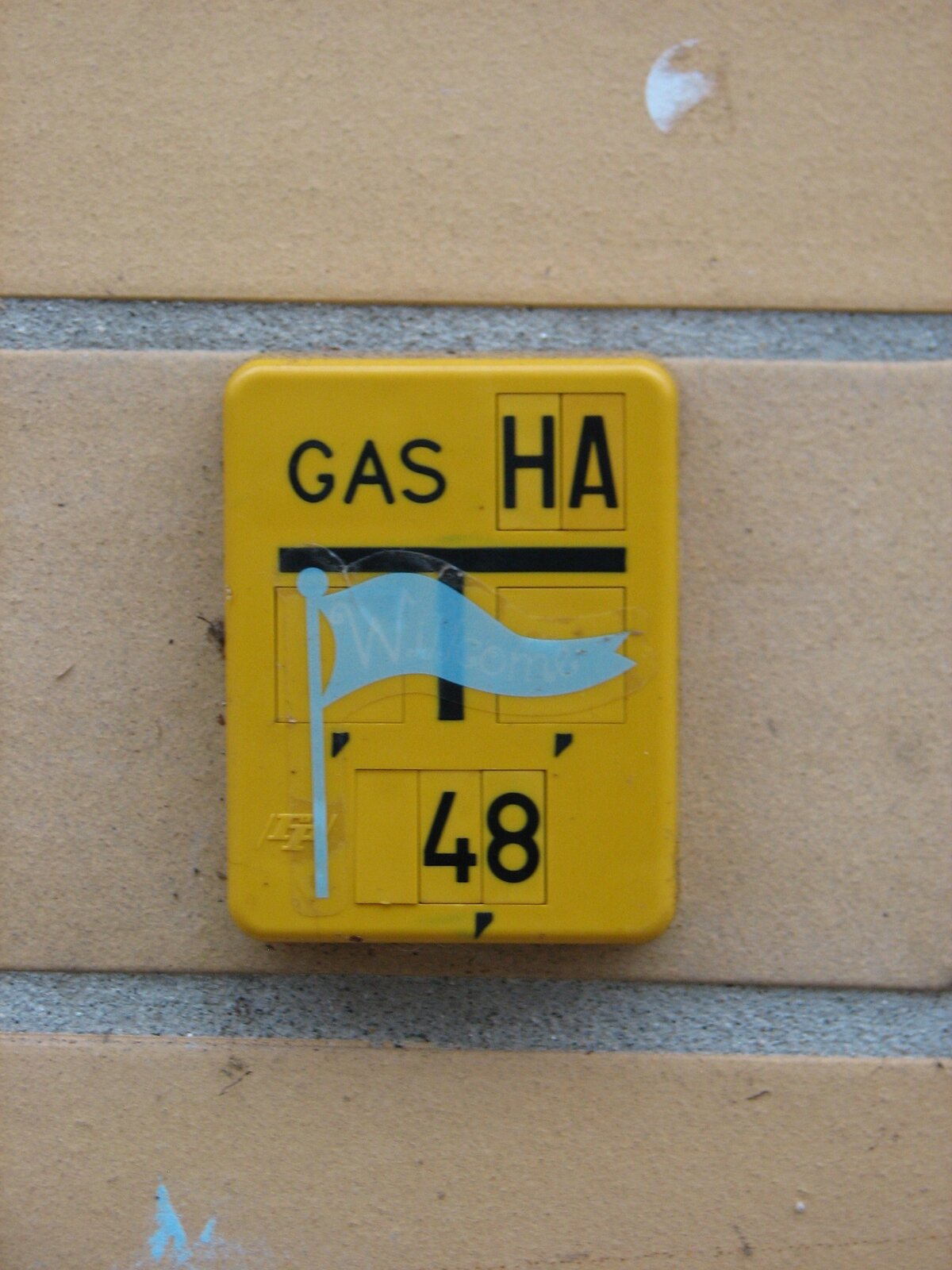 Gelbes, quadratisches Schild an einer beige gefliesten Wand mit schwarzem Text 'GAS HA' und einer stilisierten blauen Flaggenfigur, darunter die Nummer 48.