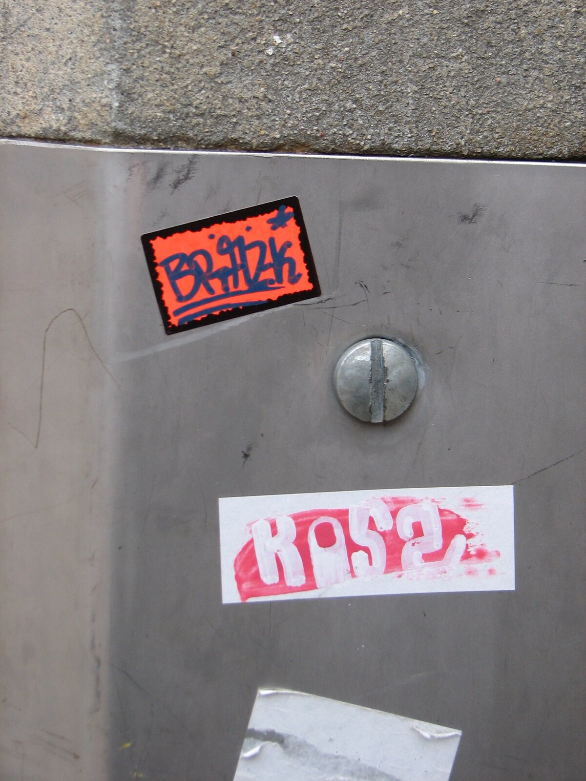 Graue Metallfläche mit zwei übereinander angebrachten Stickern in Orange und Rot, daneben ein metallischer Bolzen. Auf dem orangenen ist "BRAZK" zu lesen, auf dem rot-weißen vermutlich "KUSS".
