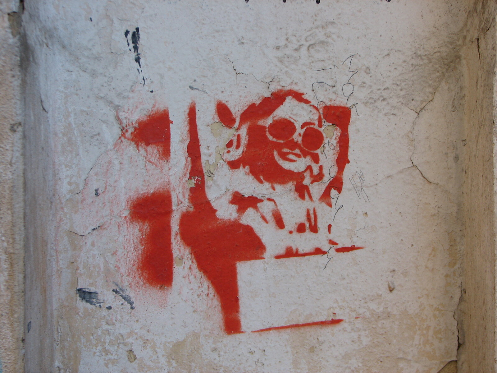 Rote Sprühfarbe auf verwitterter heller Wand: ein gestenciltes Gesicht mit markanter Sonnenbrille.