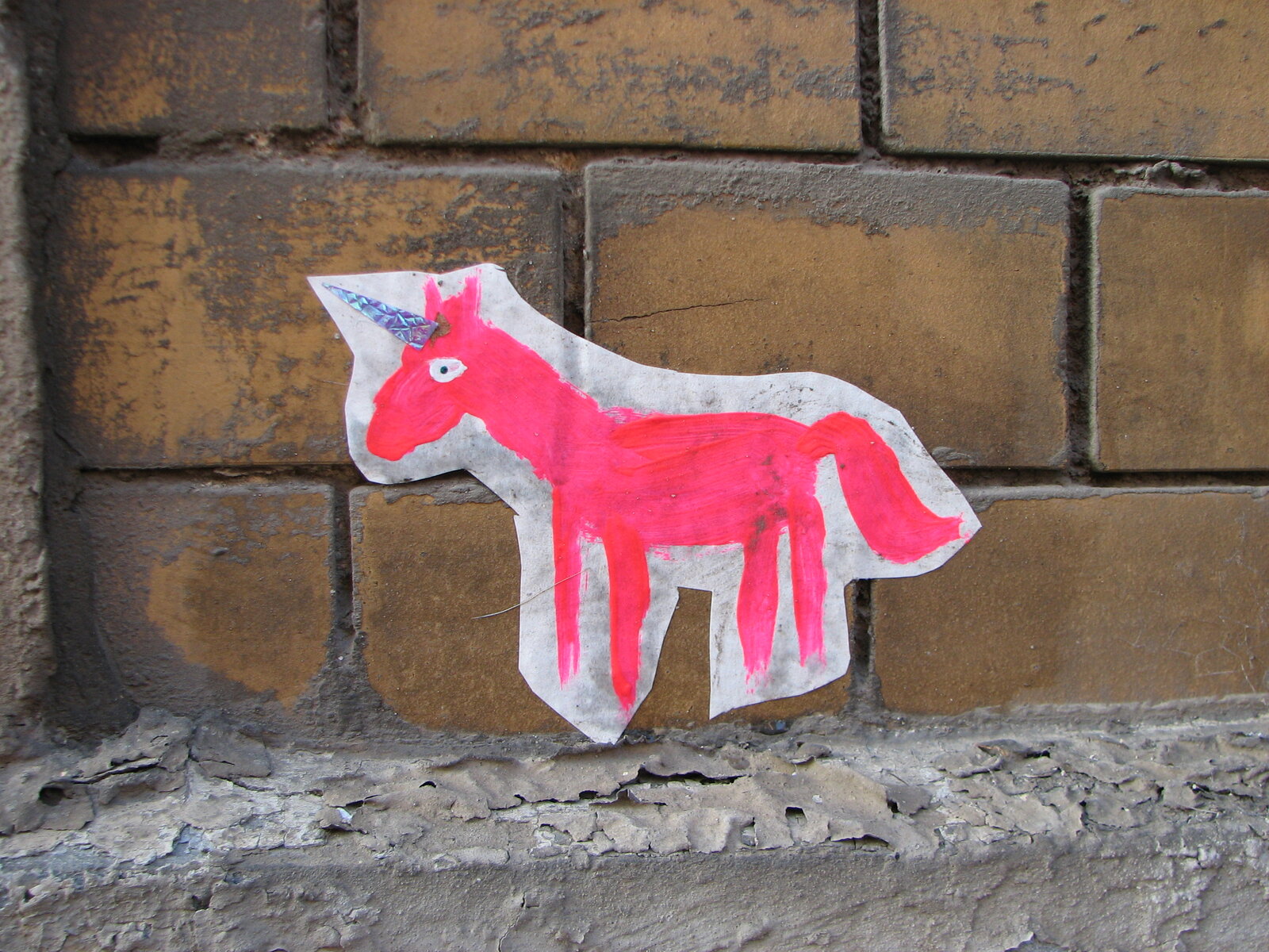 Ein rotes und weißes Einhorn-Motiv auf Papier oder Pappe, angebracht an einer Ziegelmauer. Die Figur hat einen aufgerichteten Horn, ein sichtbares Auge und ist in einfachen, klaren Formen gestaltet.