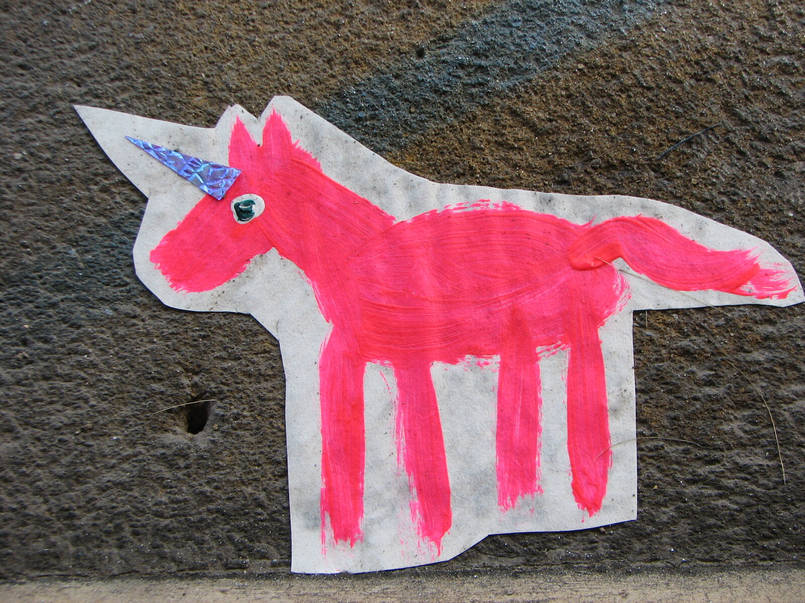Ein pinkfarbenes, schematisch gezeichnetes Einhornmotiv auf weißem Papier, mit feinen blauen Linien an Horn und Auge, an einer grauen Betonwand angebracht.