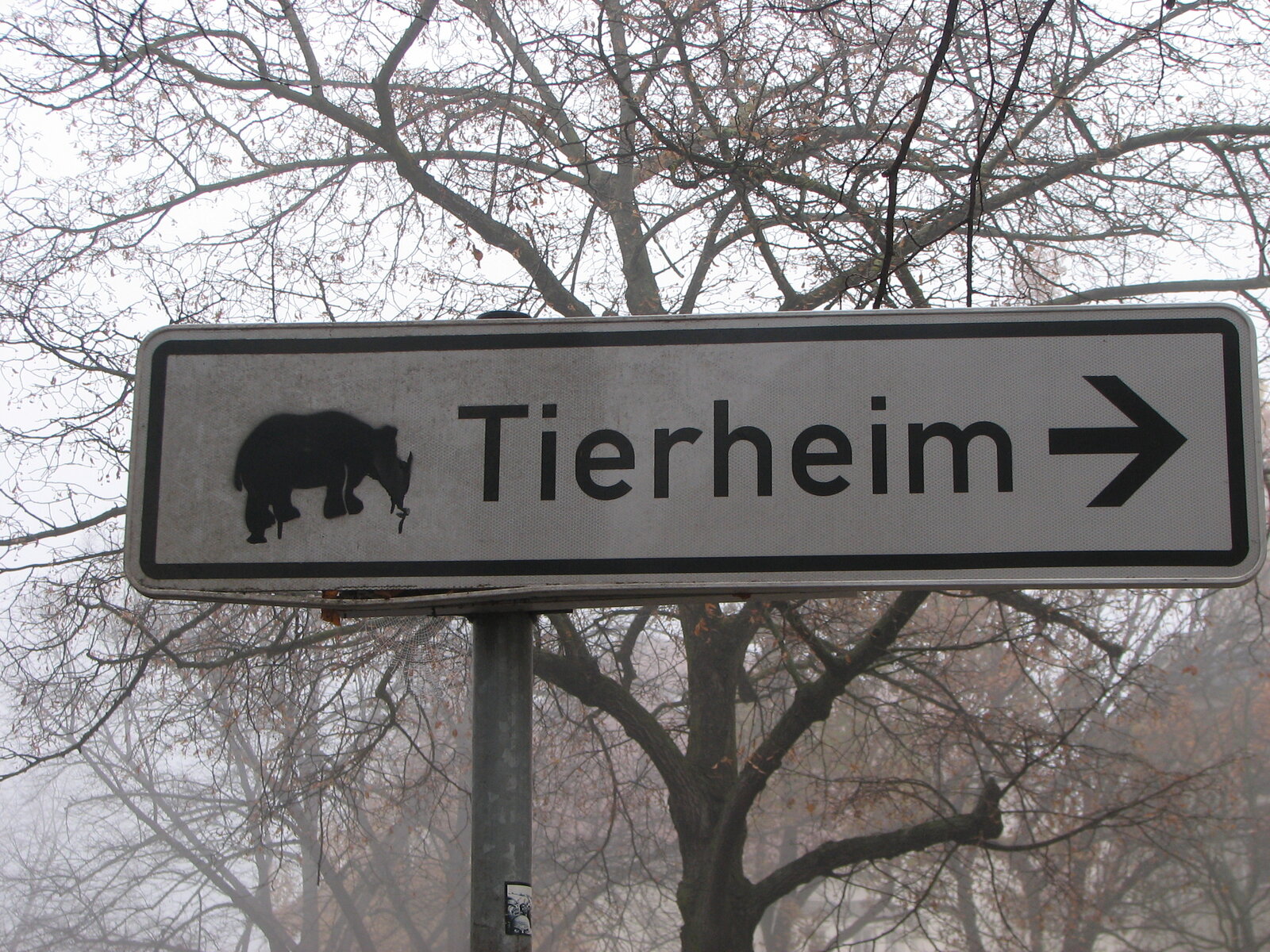 Ein Straßenschild mit Nashorn-Silhouette und der Aufschrift "Tierheim" mit Pfeil nach rechts. Im Hintergrund kahle Bäume vor grauem Himmel.