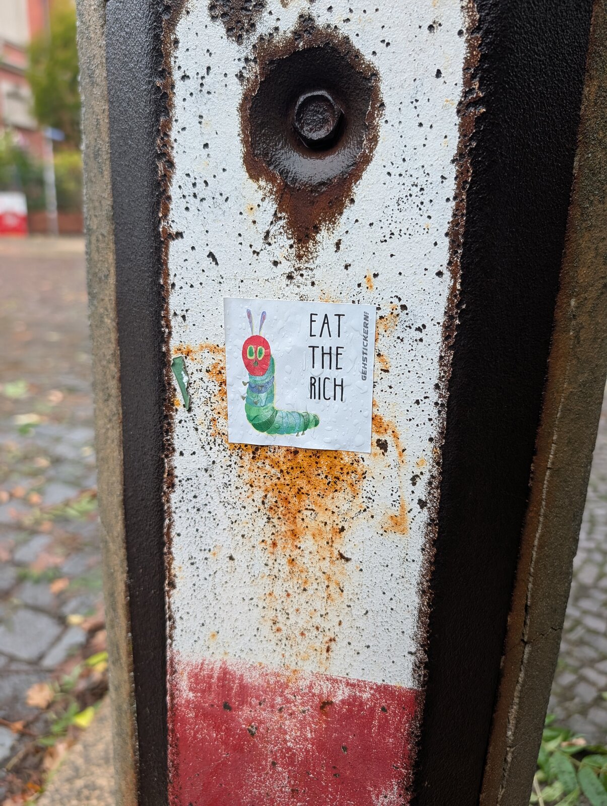 Auf einem verwitterten Betonpfeiler klebt ein rechteckiger Sticker mit der Illustration einer grünen Raupe und rotem Kopf, daneben der Text 'EAT THE RICH'.