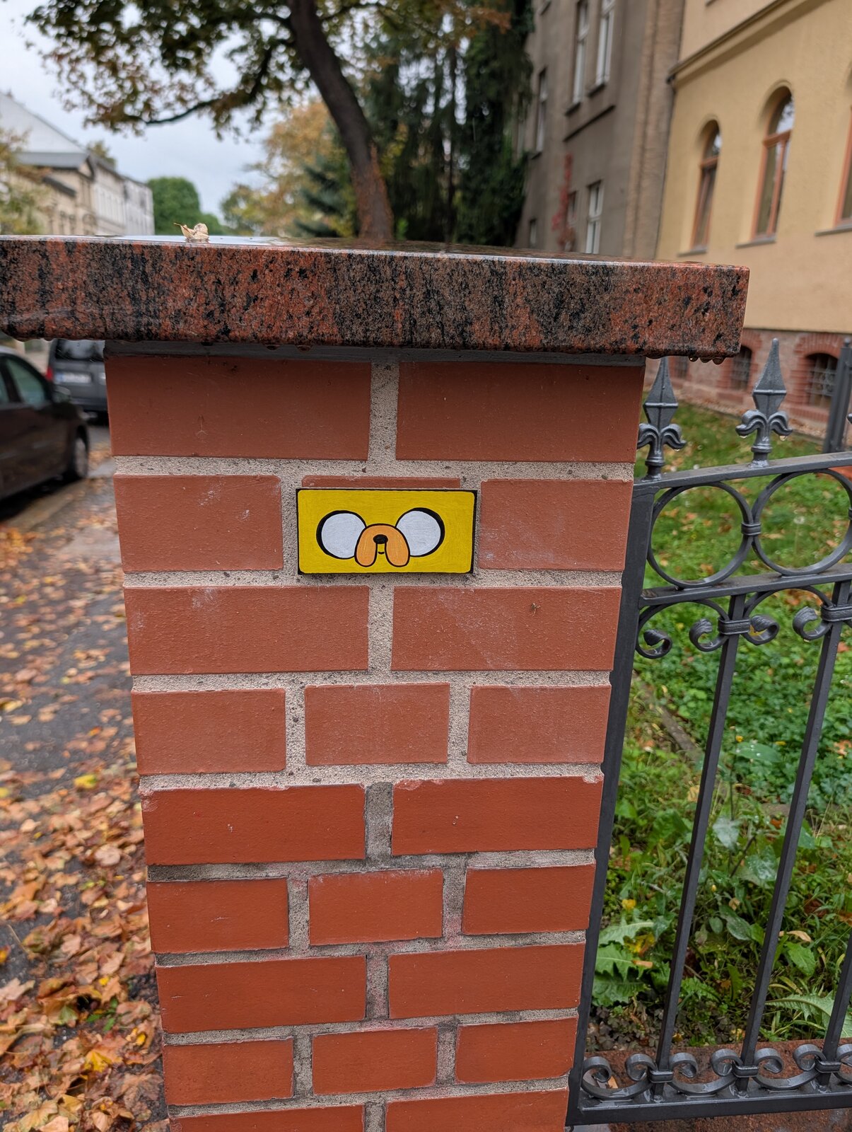 Ein kleines, gelbes Sticker-Rechteck auf roten Backsteinen einer Mauer zeigt ein einfaches Gesicht aus zwei großen weißen Kreisen als Augen und einem braunen, ovalen Mund. Abgebildet ist das Gesicht von Jake aus der Serie adventure time.