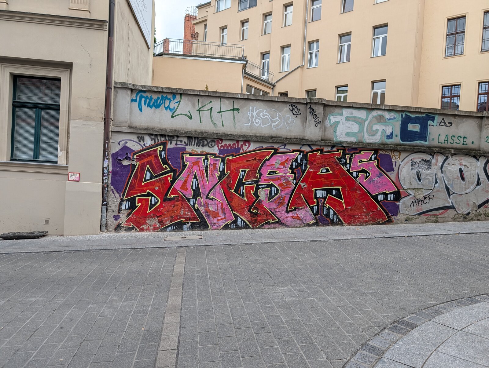 Großflächiges Graffiti an einer Mauer: Wildstyle-Schriftzug in leuchtendem Rot, Pink und Violett mit schwarzen Konturen und gelben Akzenten. Darüber und darunter mehrere überlagerte Tags in verschiedenen Farben und Stilen.