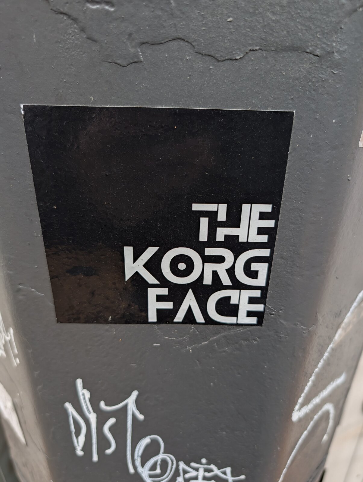 Schwarzes rechteckiges Sticker mit weißem geometrischem Schriftzug 'THE KORG FACE' auf grauer Hauswand.