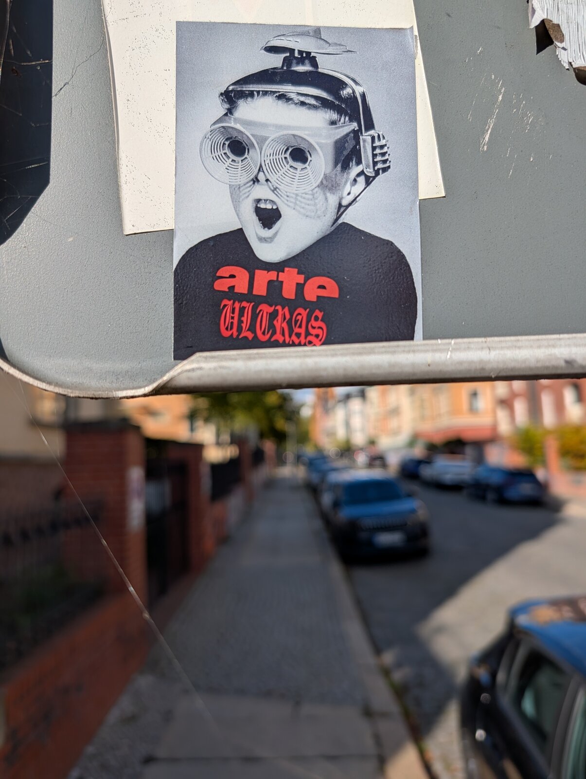 Schwarz-weißer Sticker auf Schild zeigt ein Porträt mit großen spiralförmigen Augen, Helm oder Kopfbedeckung und offenem Mund. Rote Schriftzüge 'arte' und 'ultras' befinden sich auf schwarzem Hintergrund im unteren Bereich.