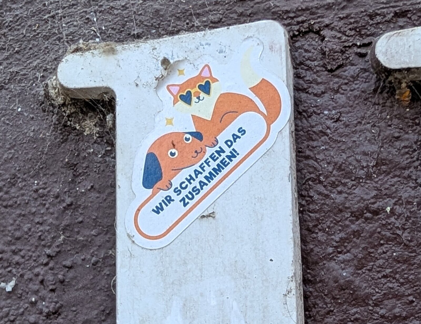 Ein Sticker auf weißem Untergrund zeigt einen braunen Fuchs in Comic-Optik mit Herzauge und rosa Ohren. Leicht versetzt darunter ist ein  orange-braun Hund zu sehen . Der Text "WIR SCHAFFEN DAS ZUSAMMEN!" verläuft darunter.
