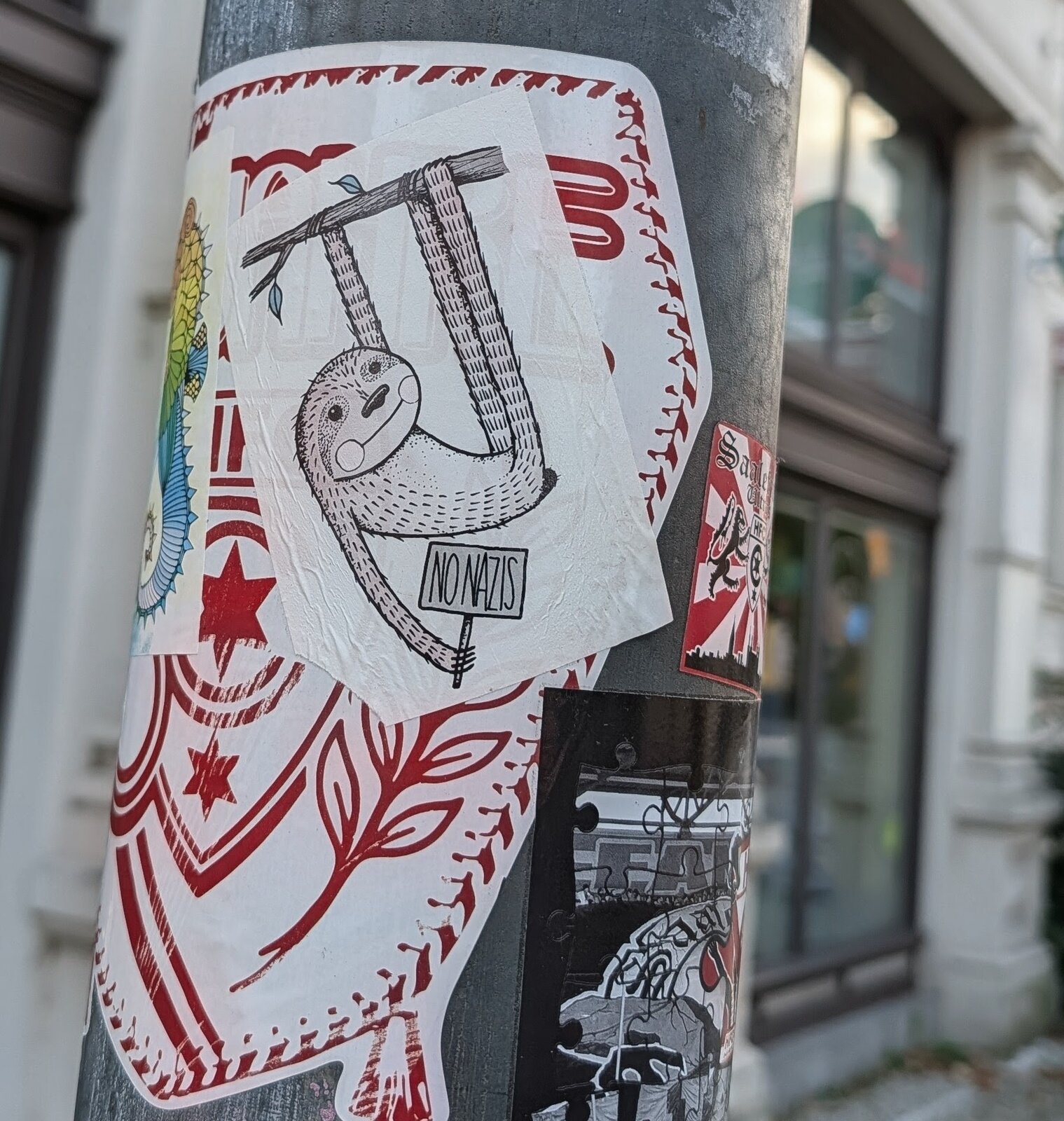 Ein Sticker auf einer grauen Metallstange zeigt eine Zeichnung eines hängenden Faultiers in Schwarz-Weiß mit der Aufschrift 'NO NAZIS'.