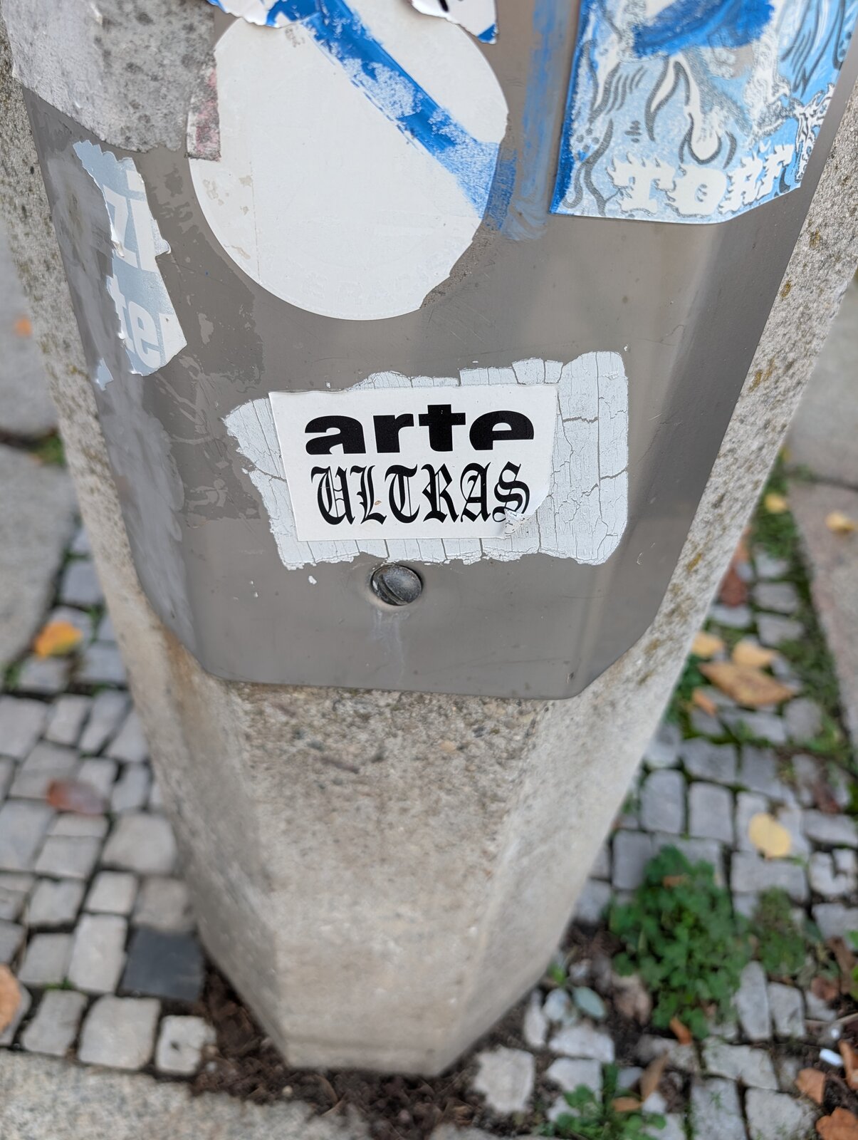 Ein weißer Sticker mit schwarzer Typografie auf einem grauen Pfosten. Der Text lautet 'arte' in serifenloser Schrift und 'ULTRAS' in verzierter Schrift.