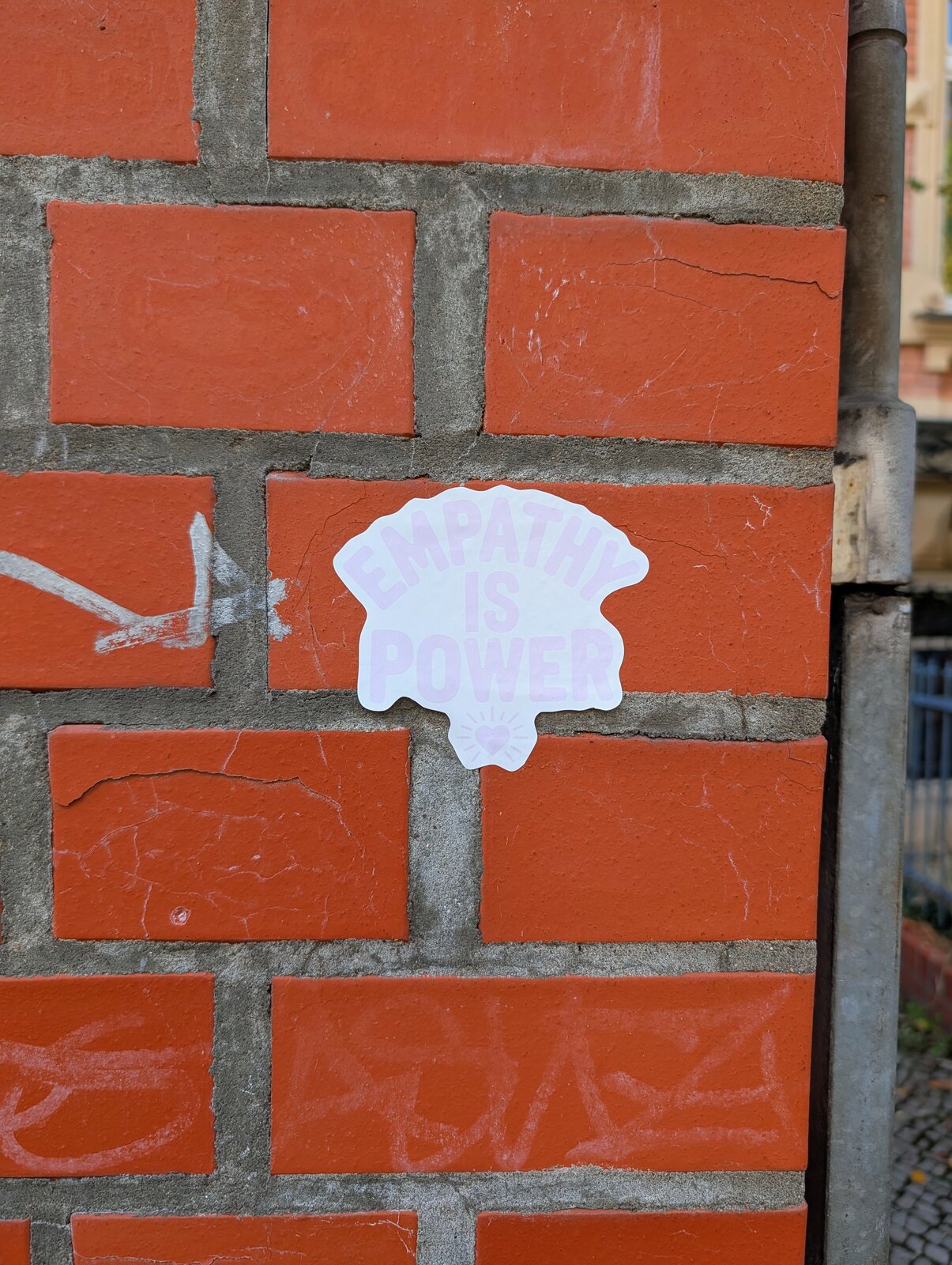 Ein weißer Sticker in Form einer abstrakten, muschelartigen Figur klebt auf einer roten Backsteinmauer mit grauem Fugenmuster. Darin steht "Empathy is power" geschrieben.