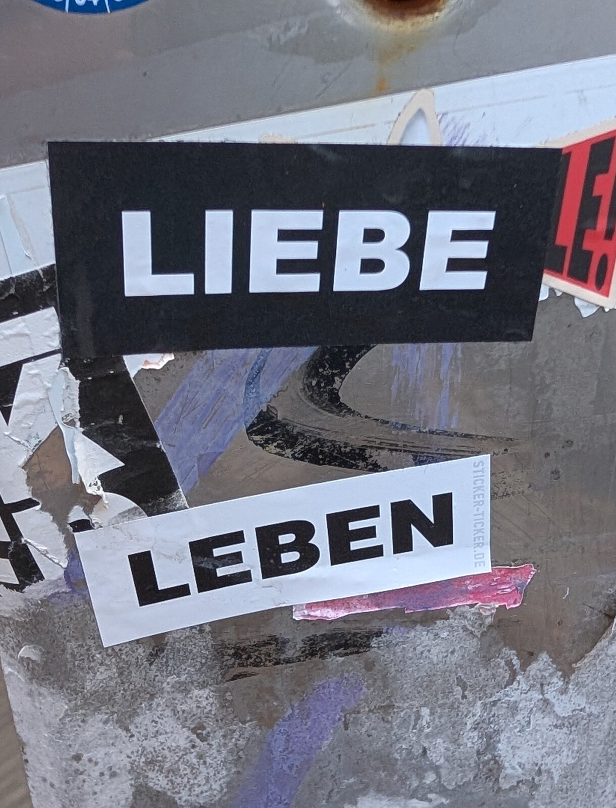 Zwei übereinandergeklebte Papier-Sticker auf verwittertem Untergrund: oben ein schwarzes Rechteck mit weißem Text "LIEBE", darunter ein weißes Schild mit schwarzem Text "LEBEN".
