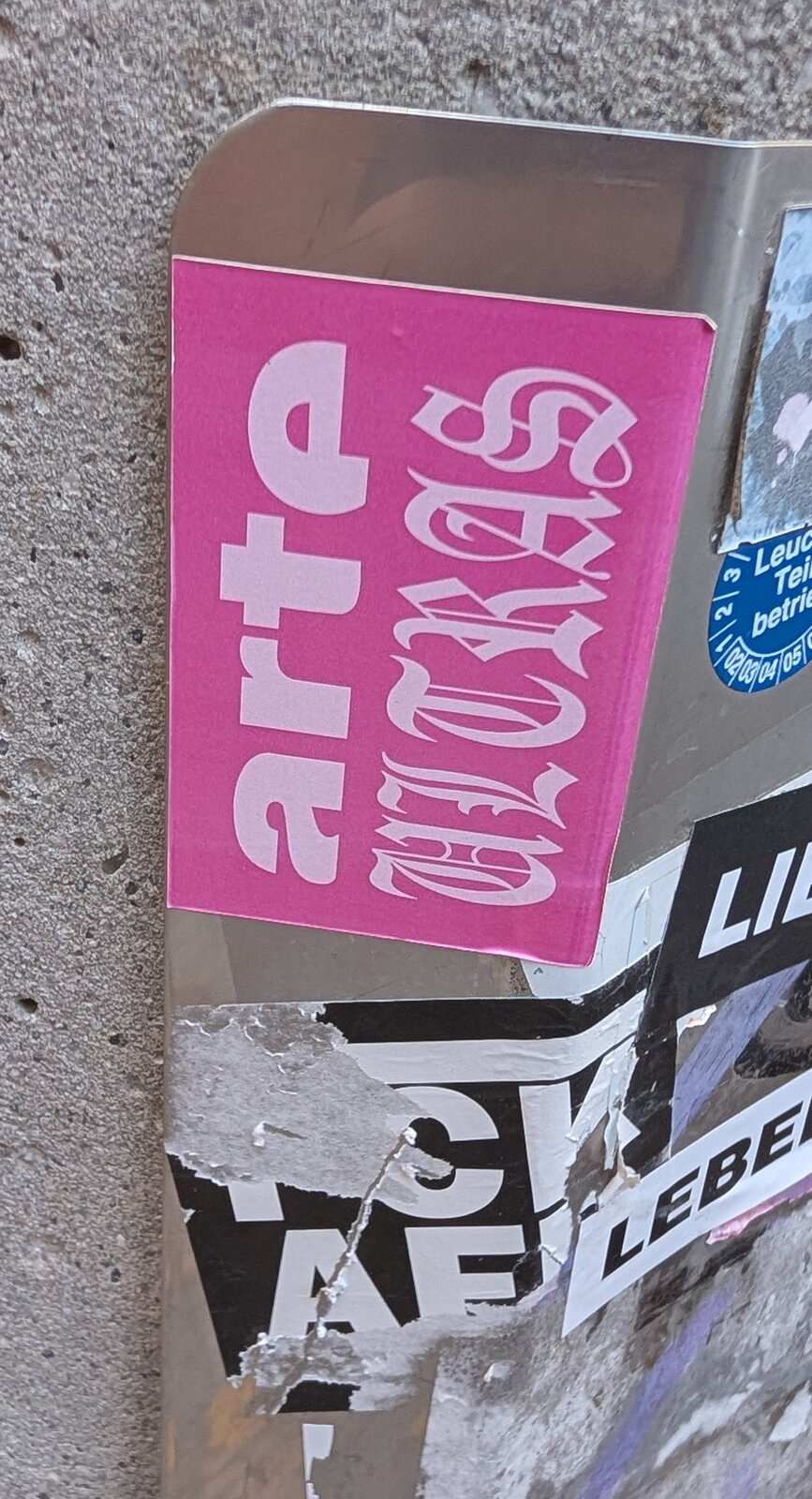 Rosafarbener Aufkleber auf einer Metallfläche mit hellrosa Schriftzügen: links in serifenloser Blockschrift "arte", rechts in einer gebrochenen, gotisch anmutenden Schrift.