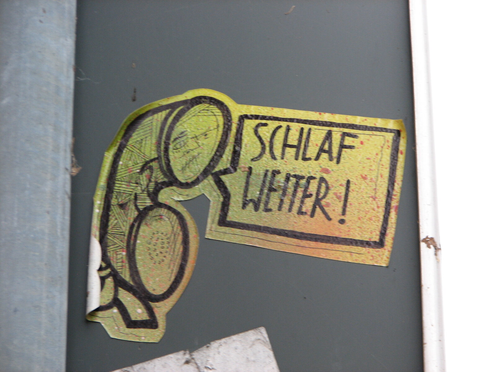 Aufkleber auf einer dunkelgrauen Metalltür: ein grün-gelb gefärbter Cartoon-Kopf im Profil mit schwarzer Konturlinie, daneben eine Sprechblase mit der Schrift „SCHLAF WEITER !“ in Großbuchstaben.