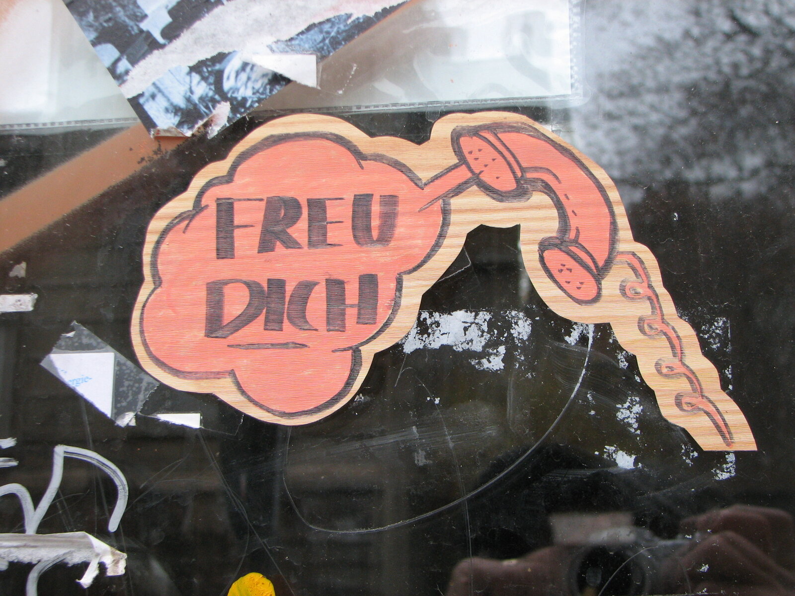 Handgemalter Sticker in Lachsrosa mit dunklem Umriss: ein Telefonhörer mit der Schrift "FREU DICH" und einer Hand, die einen Telefonhörer hält. Der Sticker ist auf eine dunkle Glasscheibe geklebt.