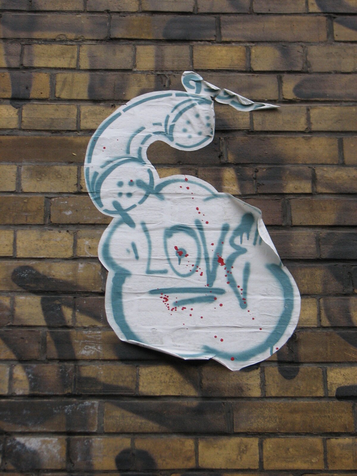 Aufgeklebtes weißes Papierplakat in Cut-out-Form an einer Ziegelwand, mit blaugrauen Sprühlinien und dem Schriftzug 'LOVE', dazu rote Farbspritzer.