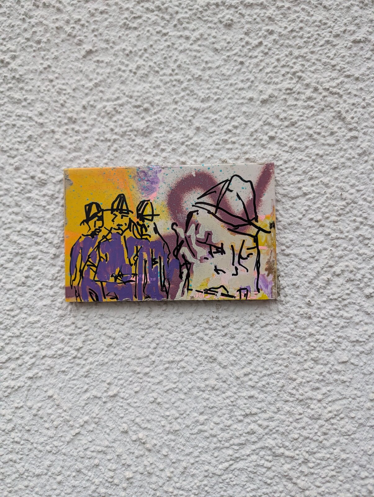 Kleine bemalte Fliese auf einer weiß strukturierten Putzwand mit schwarzen Linienzeichnungen von Figuren vor gelb-lila-farbigem Hintergrund, links drei Gestalten mit Hüten, rechts ein Kopf mit Hut auf hellem Grund.